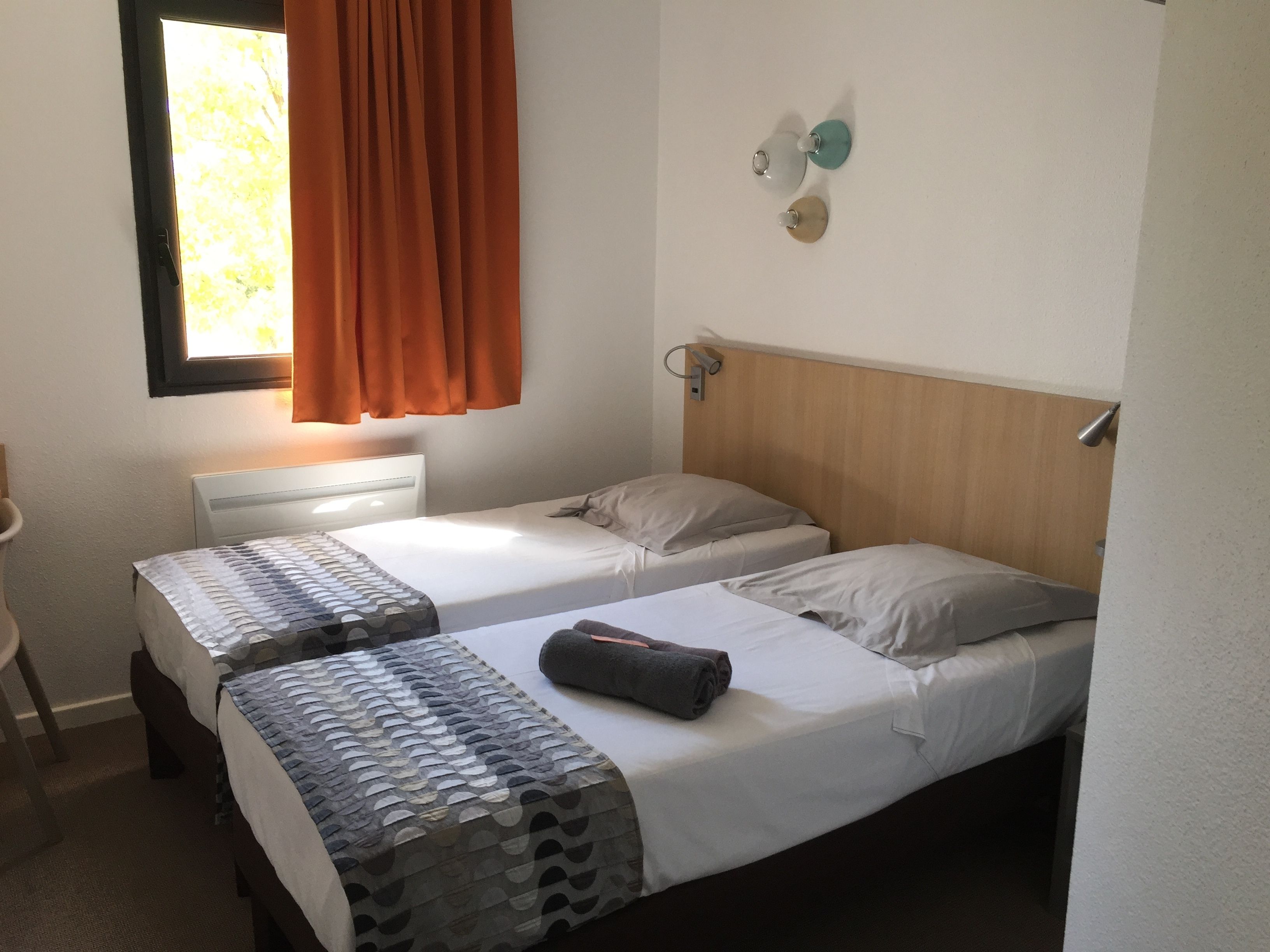 Ein Zimmer mit zwei Betten, einem Fenster mit Vorhängen, einem Nachttisch und einer Wandlampe.
