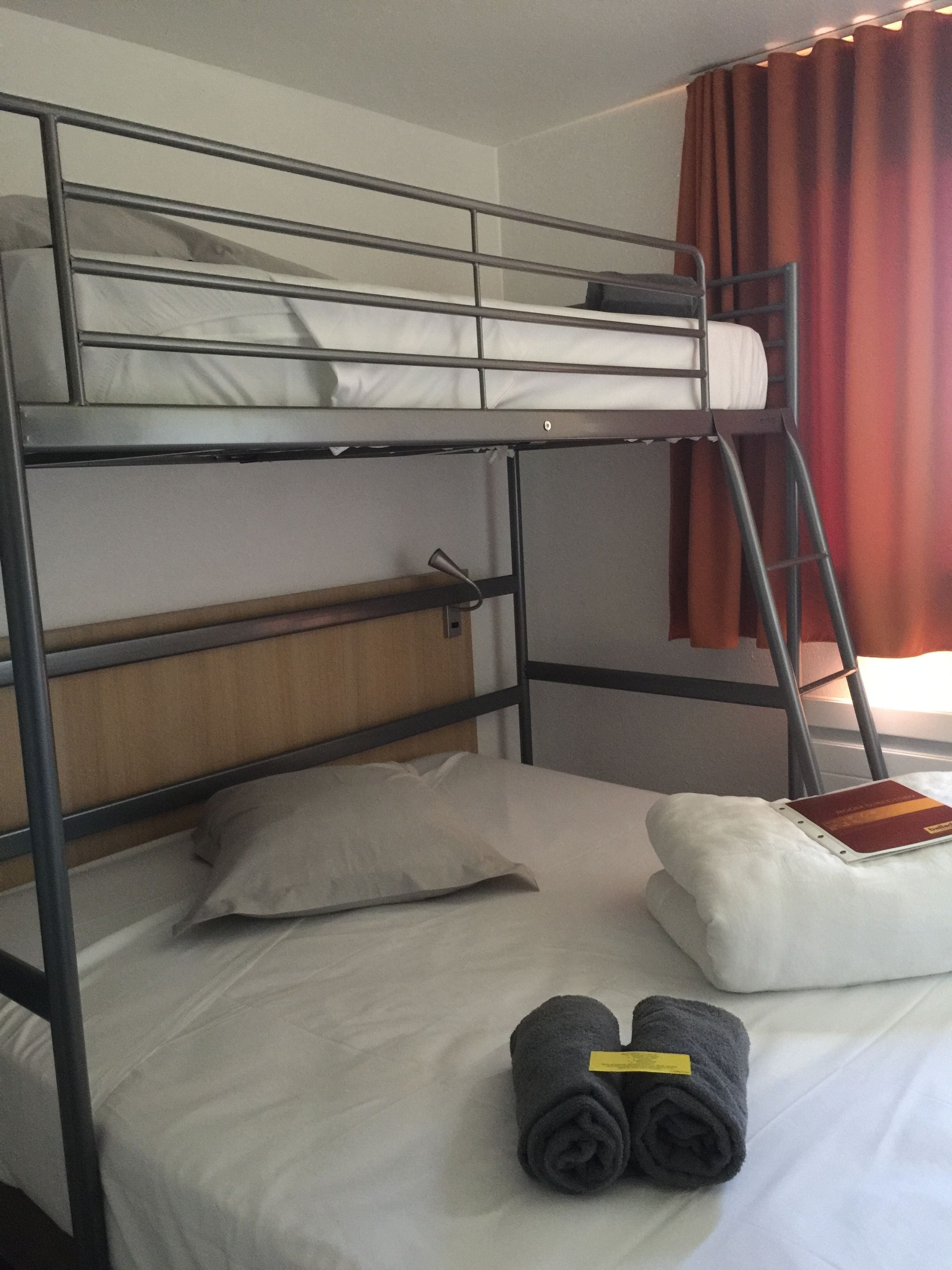A bunk bed room with a wooden headboard, a white pillow, a rolled-up towel, and a red curtain.