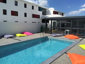 A swimming pool with colorful cushions, a glass-enclosed area, and a building with multiple windows in the background. - Kyriad Direct La Rochelle Sud Aytré (Aytre)