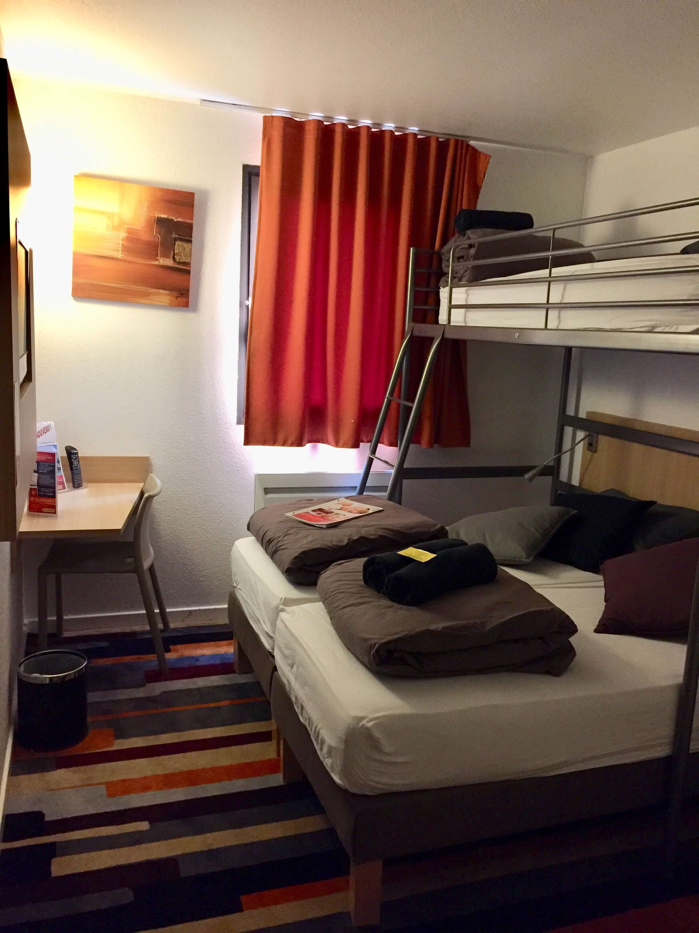 A compact hotel room with a bunk bed, a desk with a chair, a small table, and a colorful striped rug.