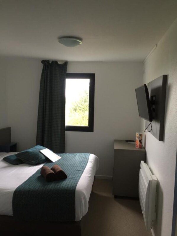 A hotel room with a bed, a small bedside table, a television, and a window with curtains.