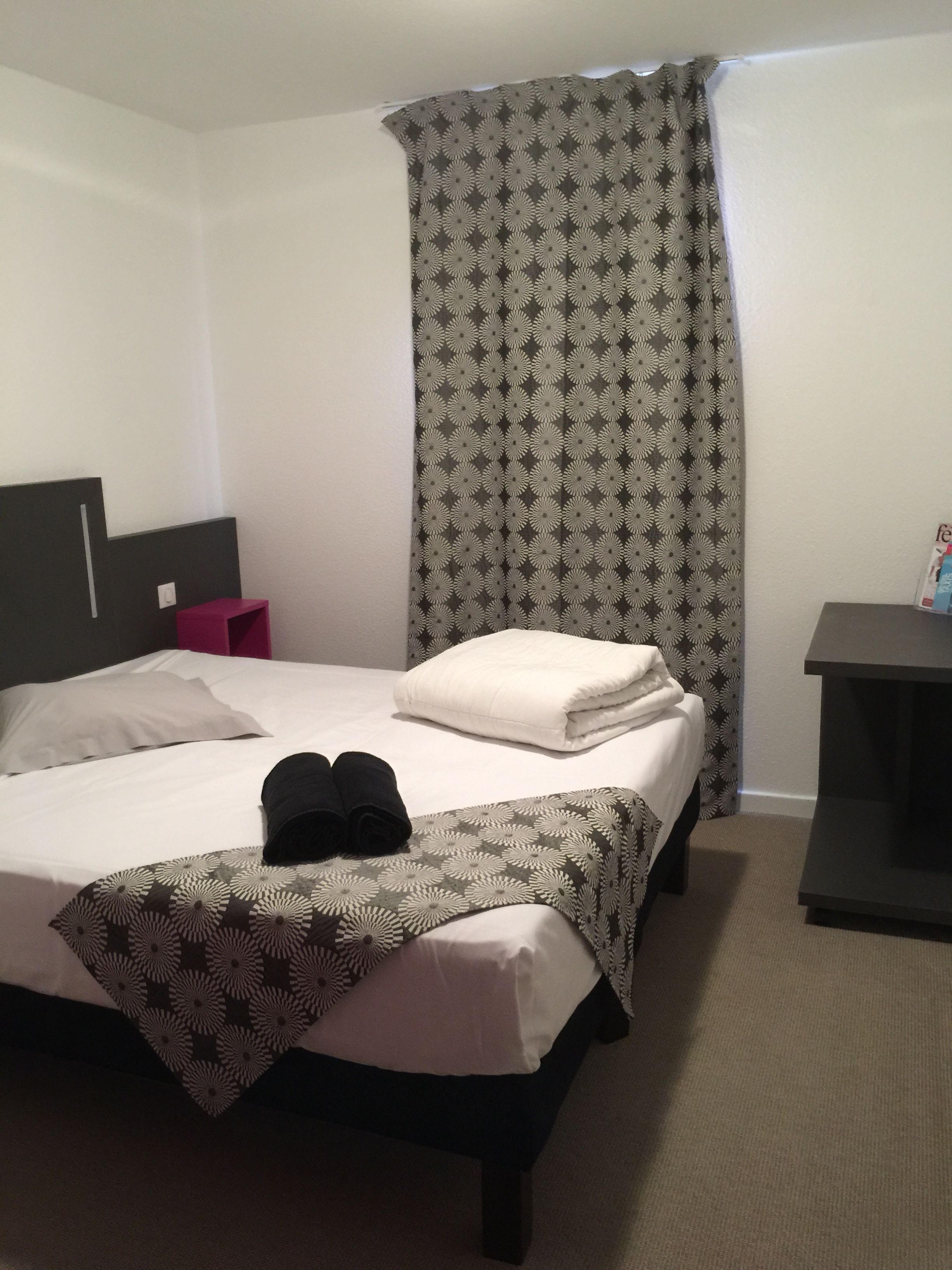 Una camera d'albergo con un letto rifatto, un comodino, una sedia e una tenda.