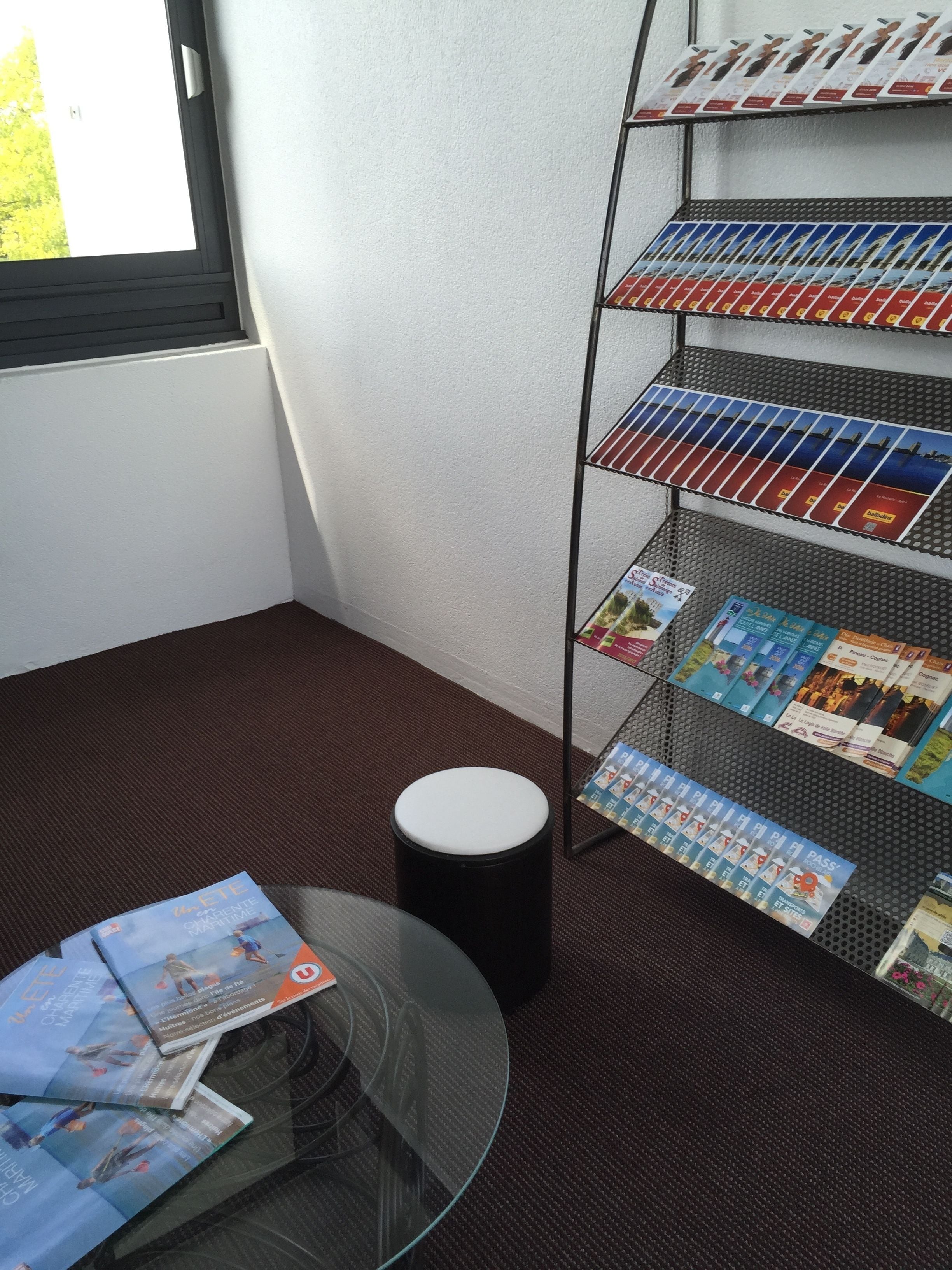 A room with a glass table displaying brochures, a black cylindrical object, and a metal rack with brochures.