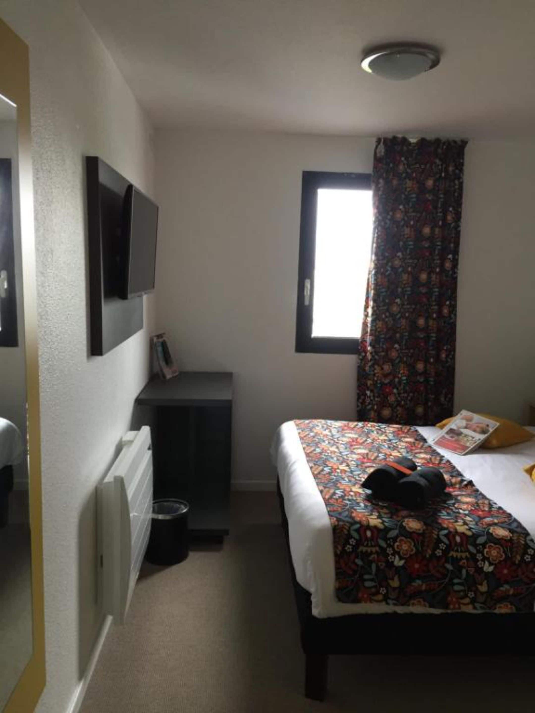 A bedroom with a bed, a TV mounted on the wall, a window with a curtain, and a radiator.