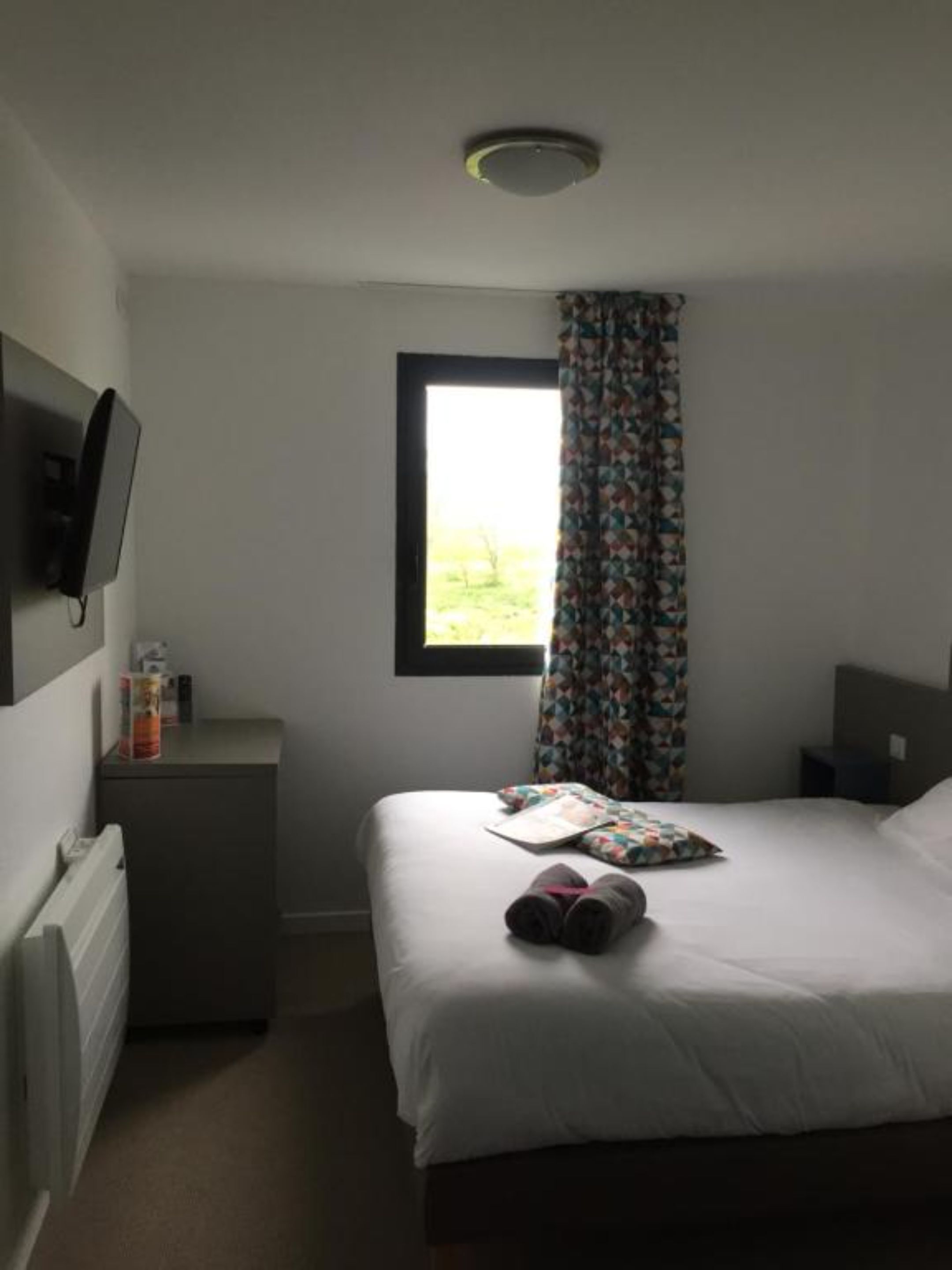A small, simple hotel room with a bed, a bedside table, a TV, and a window with a curtain.