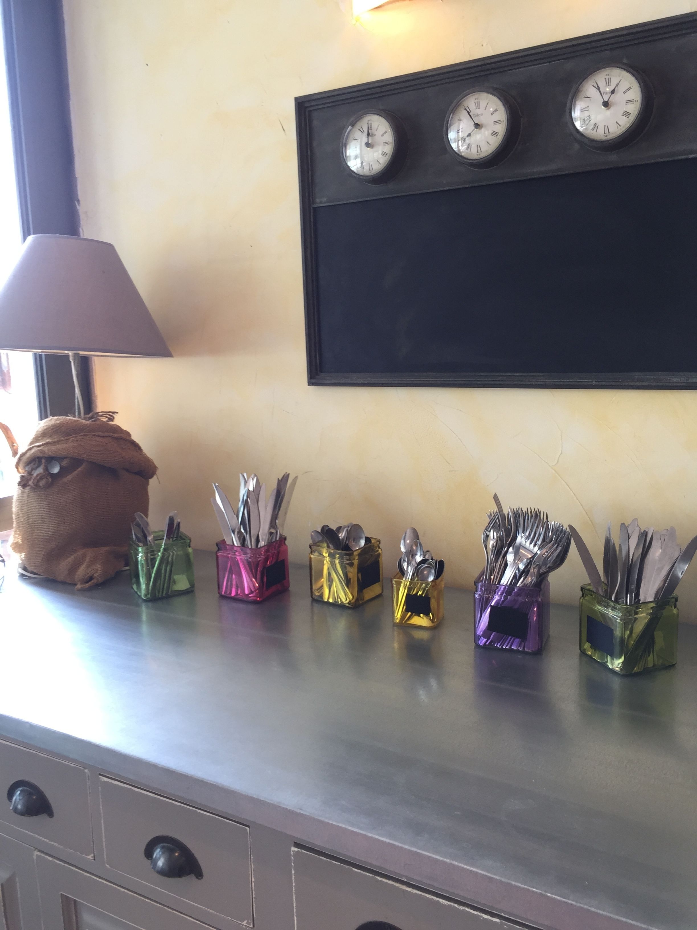 Una encimera de cocina con cuatro relojes de pared y cinco recipientes de vidrio que contienen utensilios.
