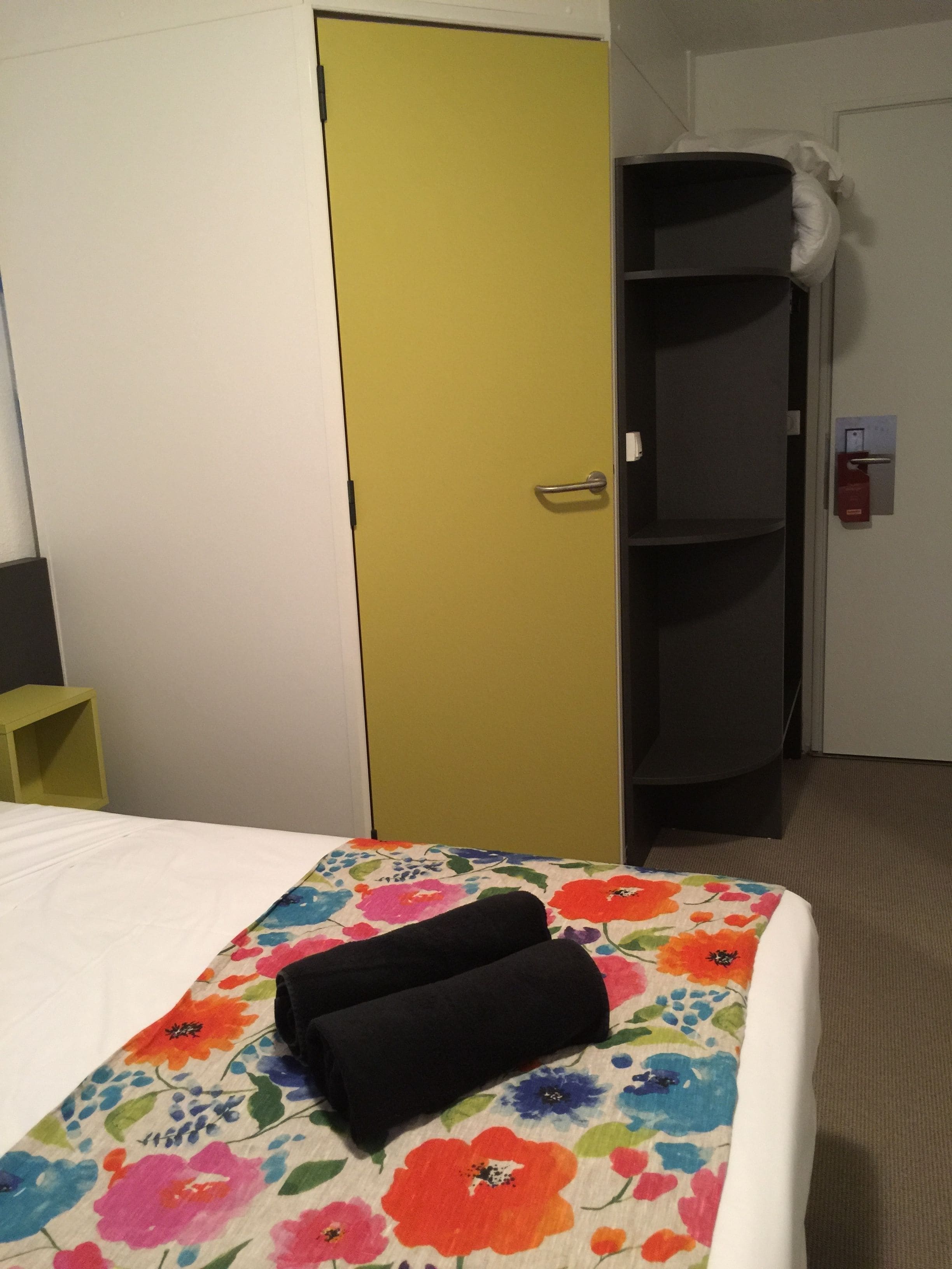 A hotel room with a bed, a yellow door, a black wardrobe, and a white wall.