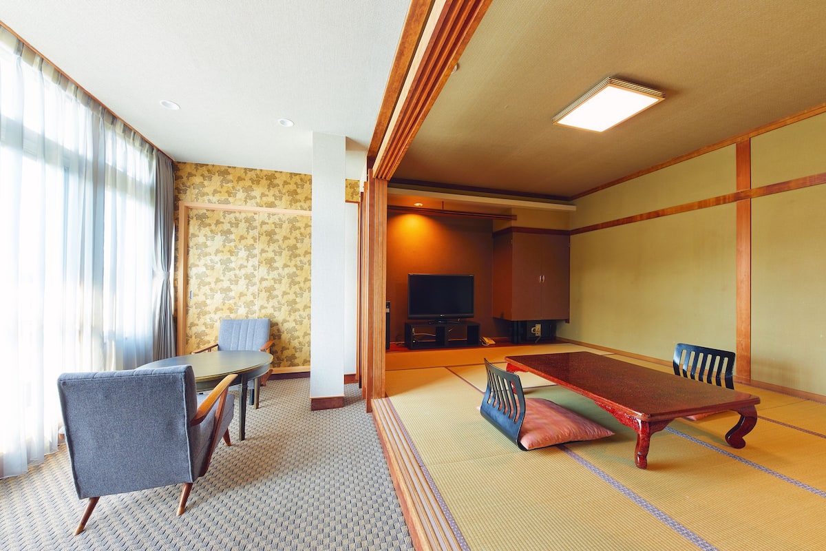 A traditional Japanese-style room with tatami flooring, a low table with cushions, a television, and a sofa.
