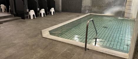A swimming pool with a tiled floor and a metal ladder. There are black and white stools and a wall-mounted TV.