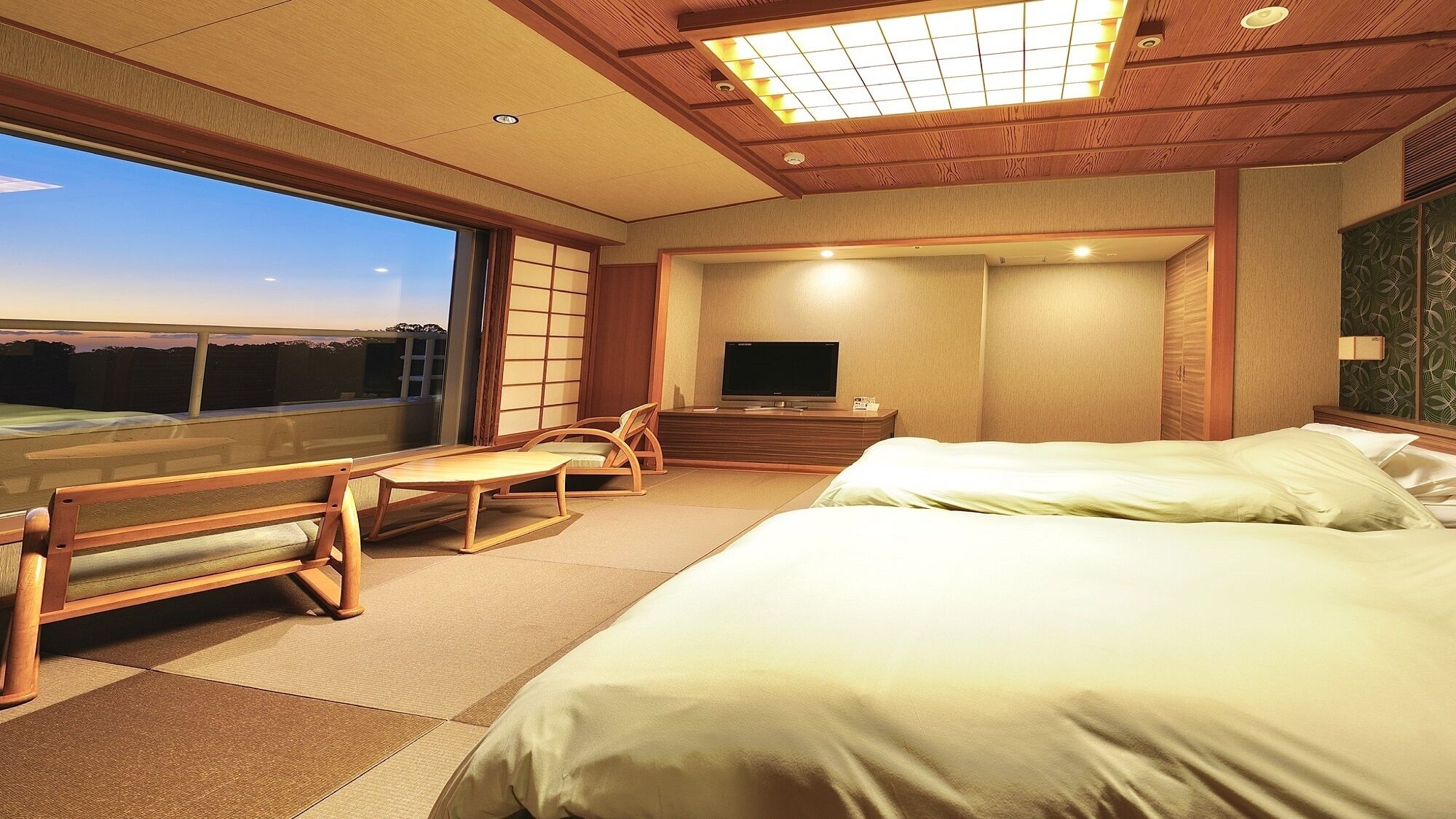 Quarto de hotel com cama, televisão, área de estar com mesa e cadeiras, e vista através de uma janela grande.