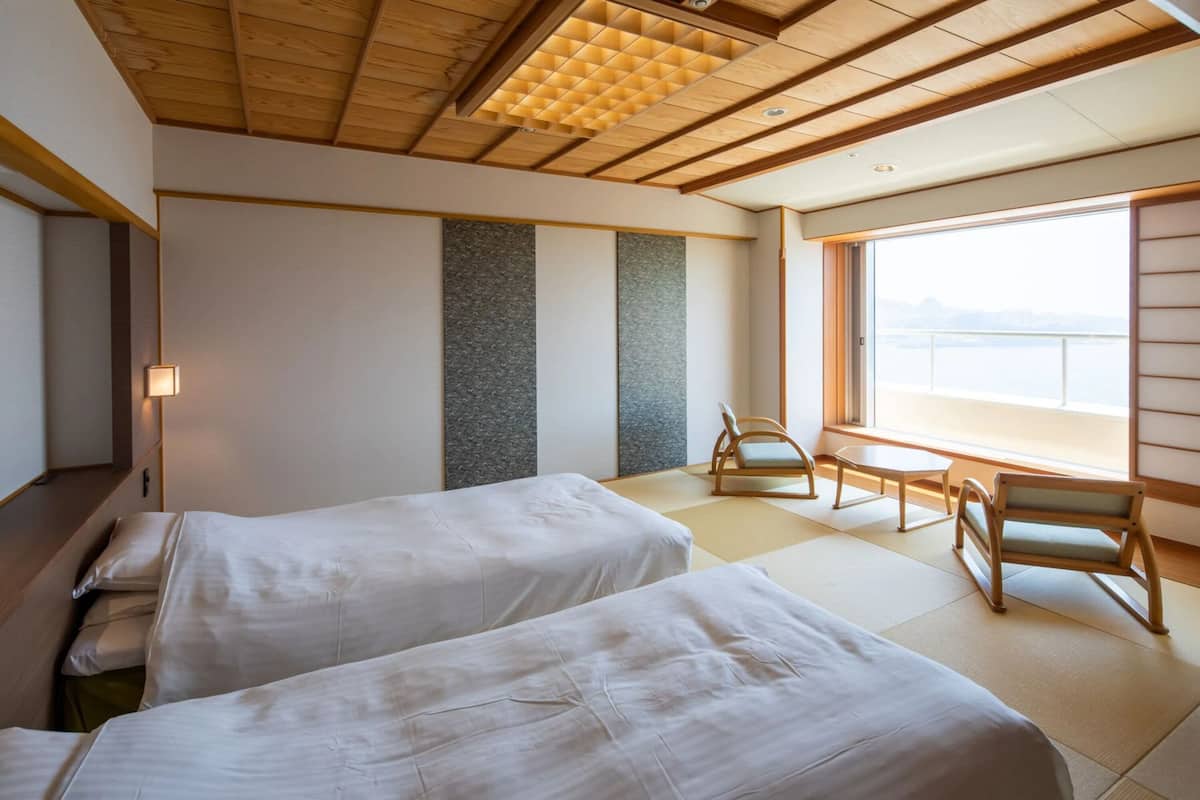A hotel room with two beds, a wooden ceiling, a large window with a view, tatami flooring, and a traditional Japanese sliding door.
