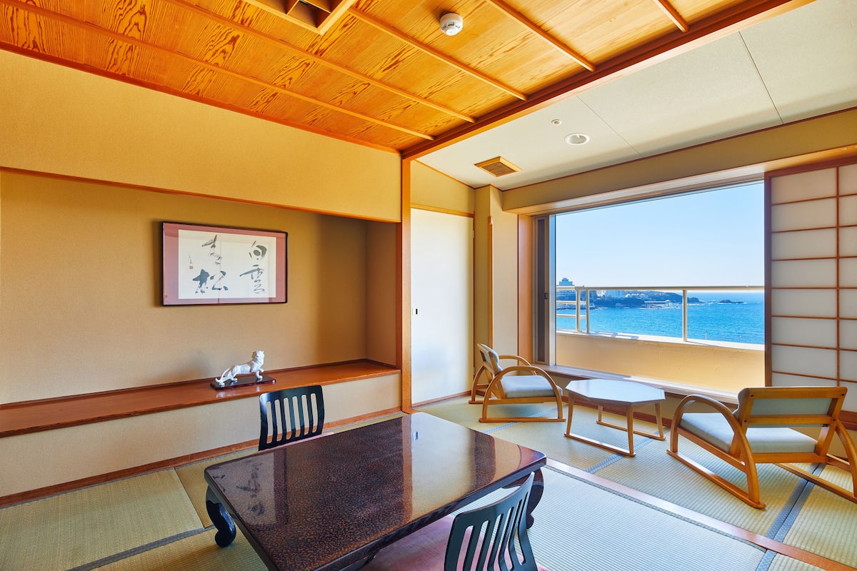 A traditional Japanese room with tatami flooring, a wooden table, chairs, a framed artwork, and a view of the sea through large windows.