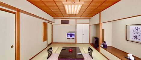 Um quarto japonês tradicional com piso de tatame, uma mesa baixa com cadeiras, uma televisão e uma obra de arte emoldurada na parede.