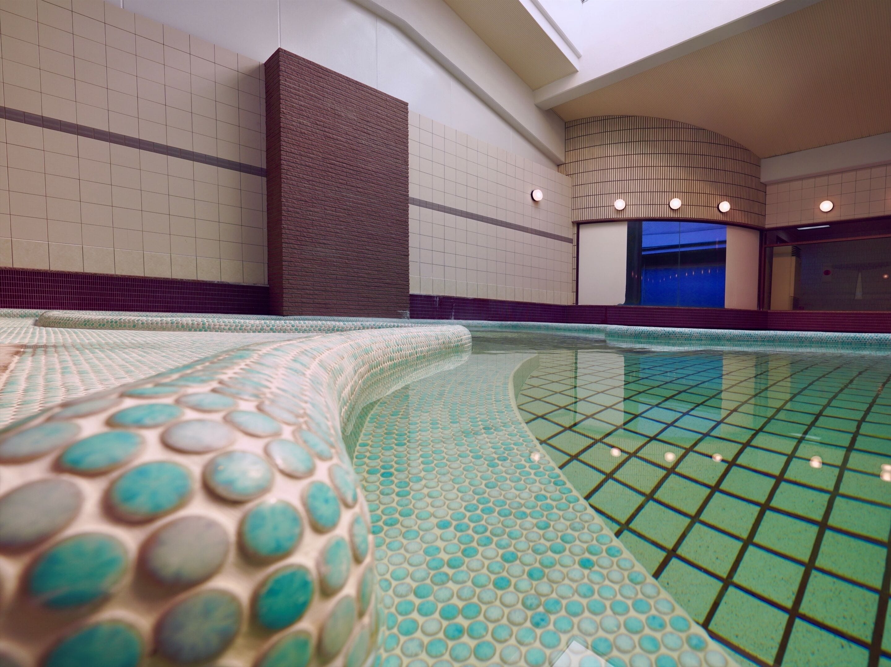 An indoor swimming pool with a tiled floor and walls, a curved edge, and a ceiling with recessed lighting.