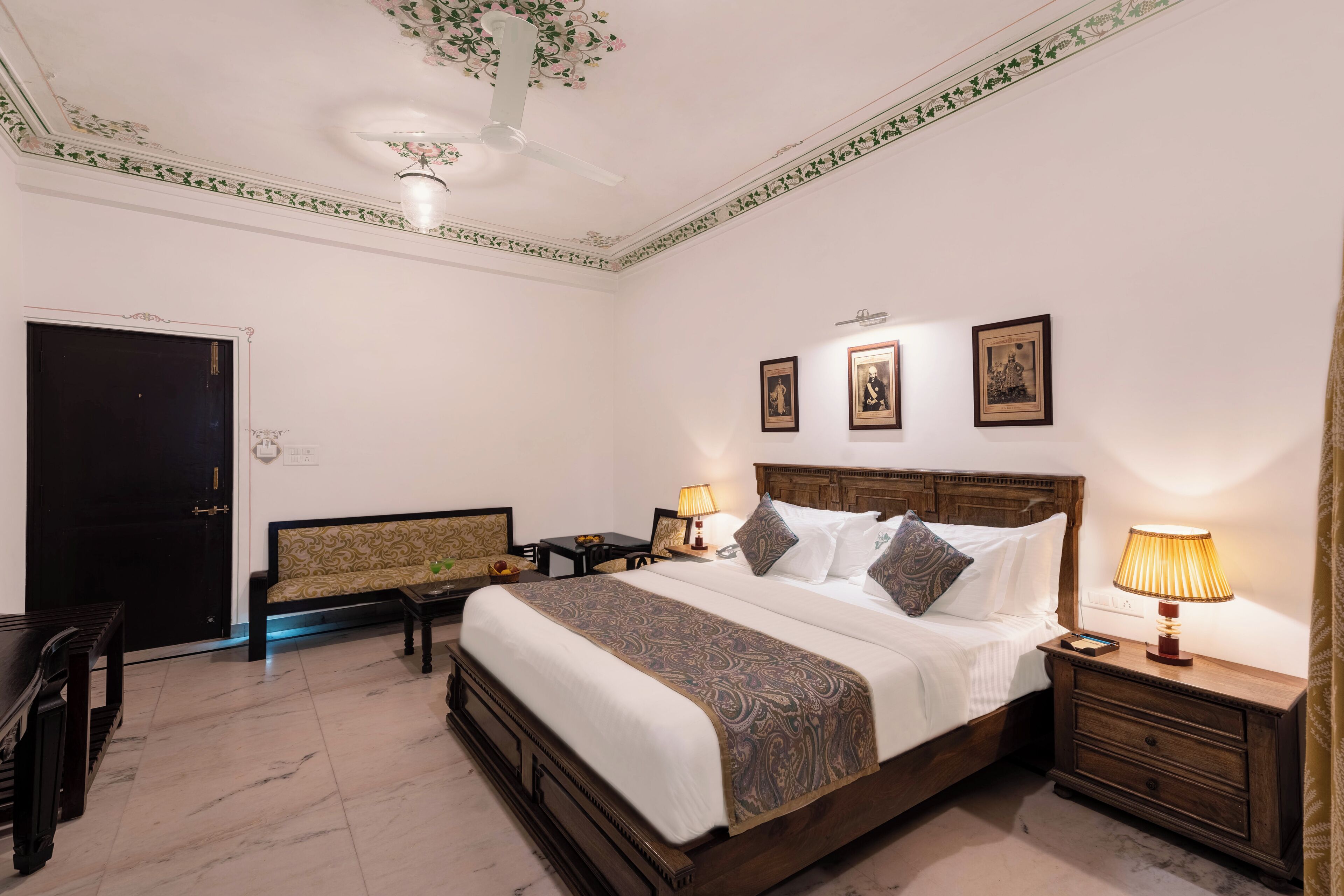 Una habitación de hotel con una cama grande, dos mesitas de noche con lámparas, un sofá, un banco de madera y una puerta.