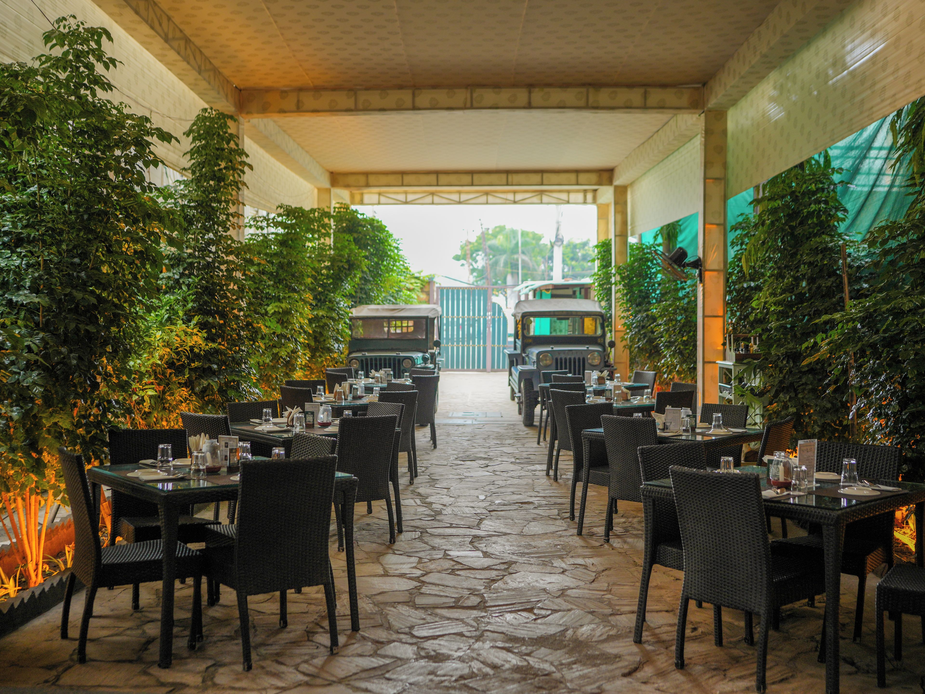 Un restaurant en plein air avec un sol en pierre, des tables et des chaises en osier, et une jeep visible au fond.
