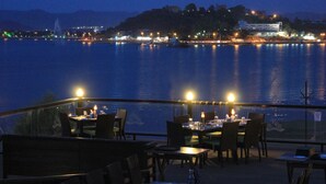 An outdoor dining area with tables and chairs, overlooking a body of water with city lights in the background. - Panna Vilas - A Lake Facing Boutique Hotel (Udaipur)