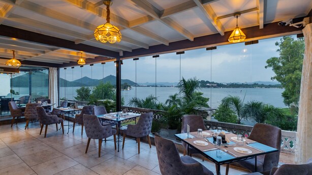 A restaurant with tables and chairs, large windows, and a view of a body of water. - Panna Vilas - A Lake Facing Boutique Hotel (Udaipur)