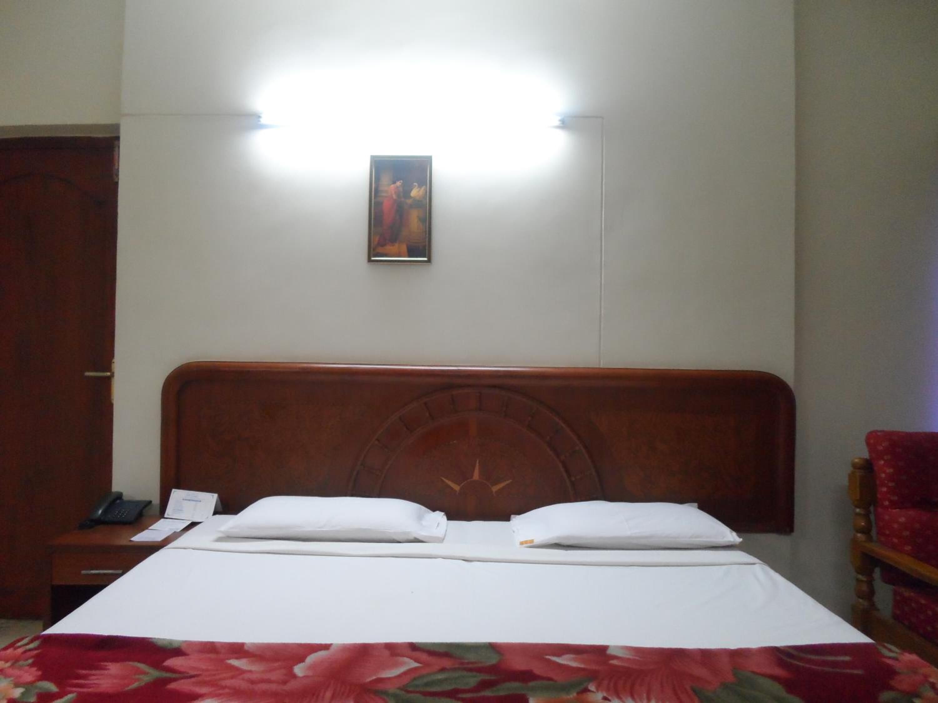 A hotel room with a wooden headboard, a bed with white linens, a floral-patterned bedspread, a nightstand with a phone and papers, and a framed picture on the wall.