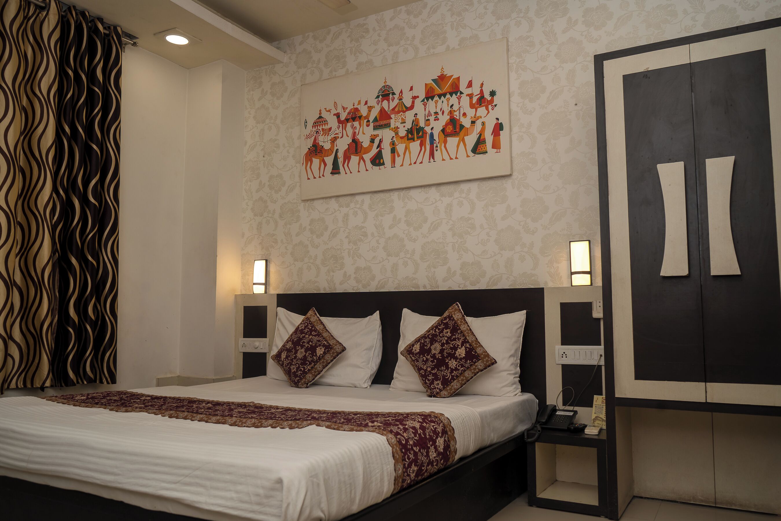 A neatly made bed with a patterned bedspread, two decorative pillows, a wall-mounted artwork depicting a scene with camels, and a wardrobe with a unique design.