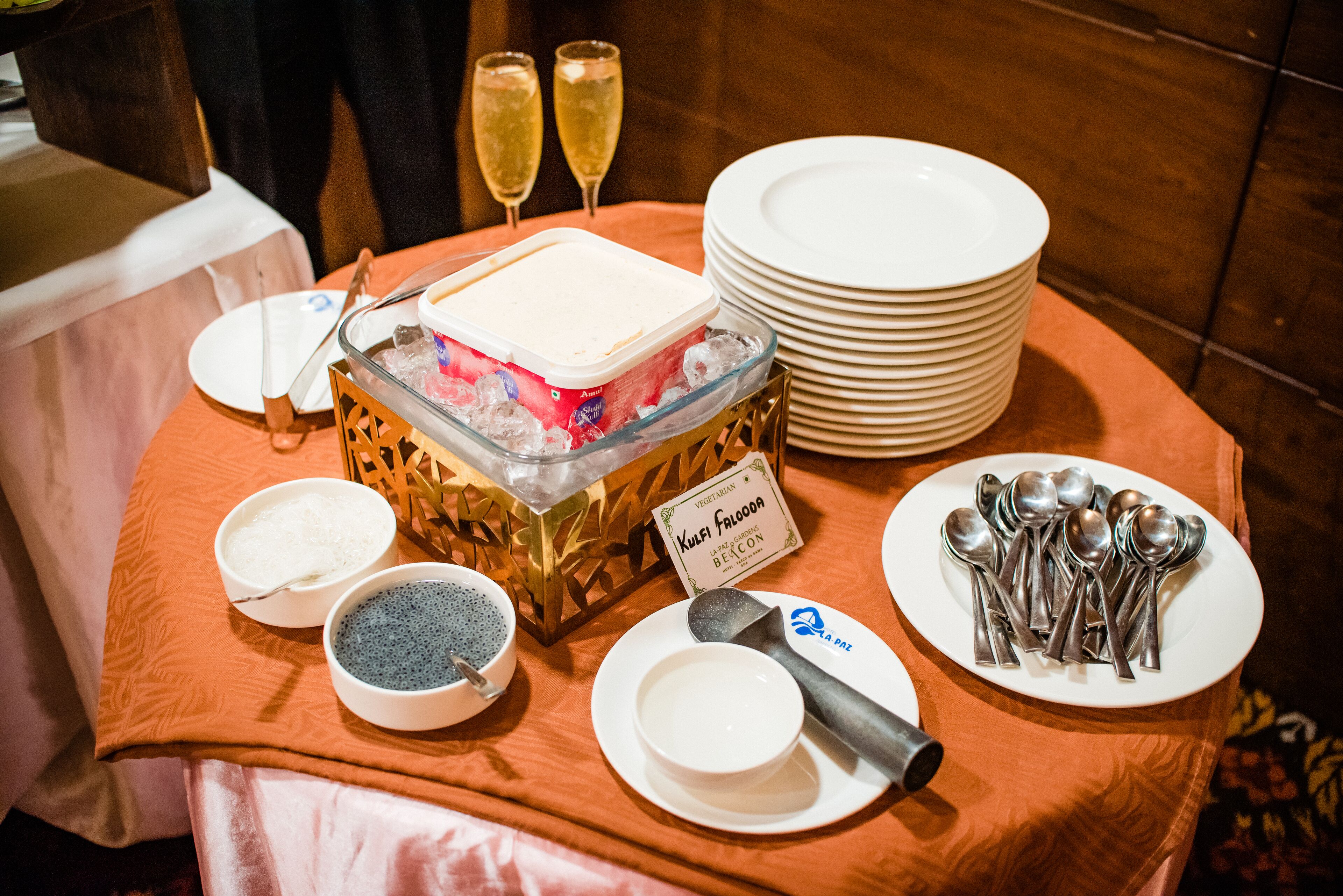 A buffet table with various food items, a sign for Kufi Falooda, and a stack of white plates and spoons.