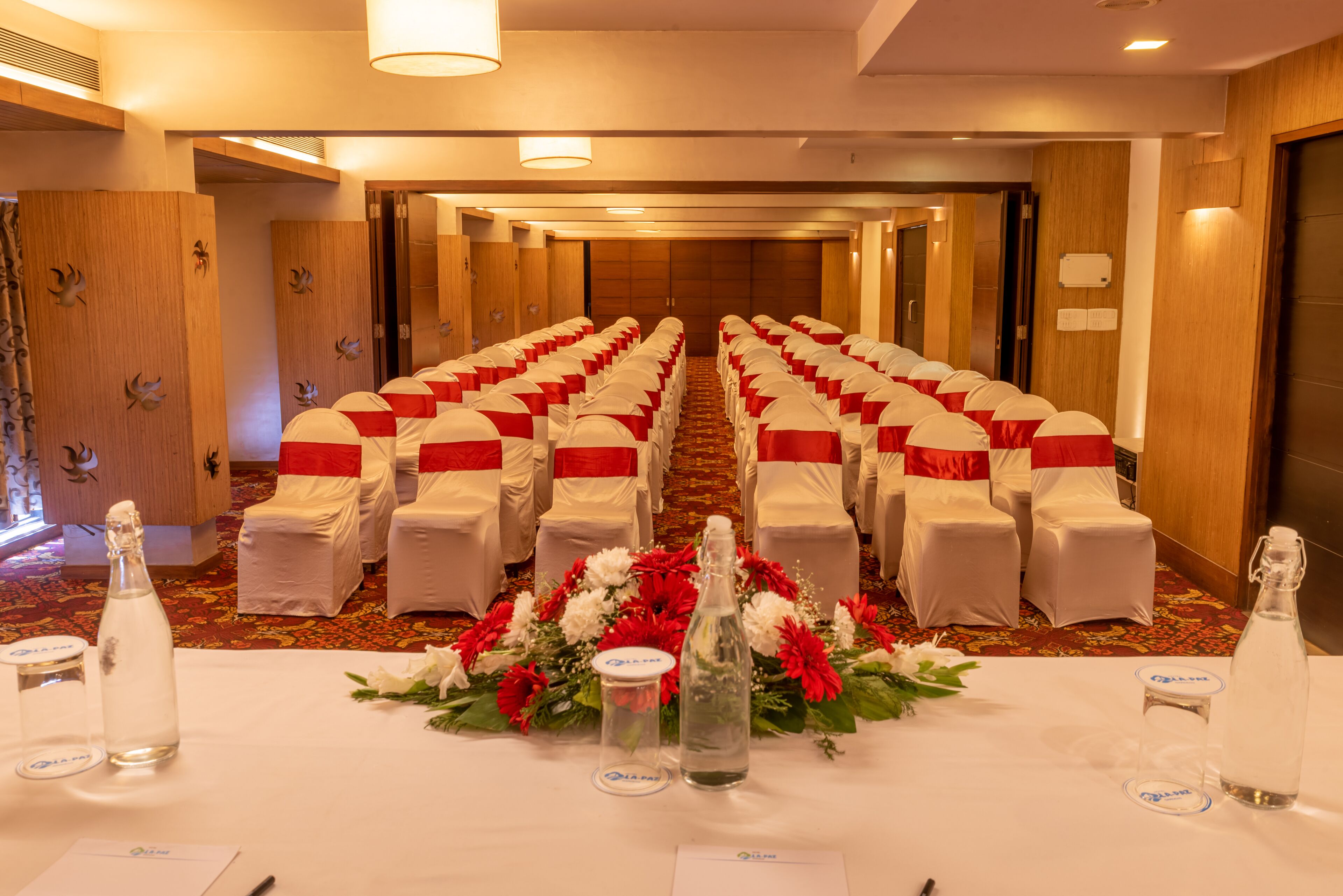 Ein Hotelkonferenzraum mit Reihen weißer Stühle, roten Bändern und einer floralen Tischdekoration.