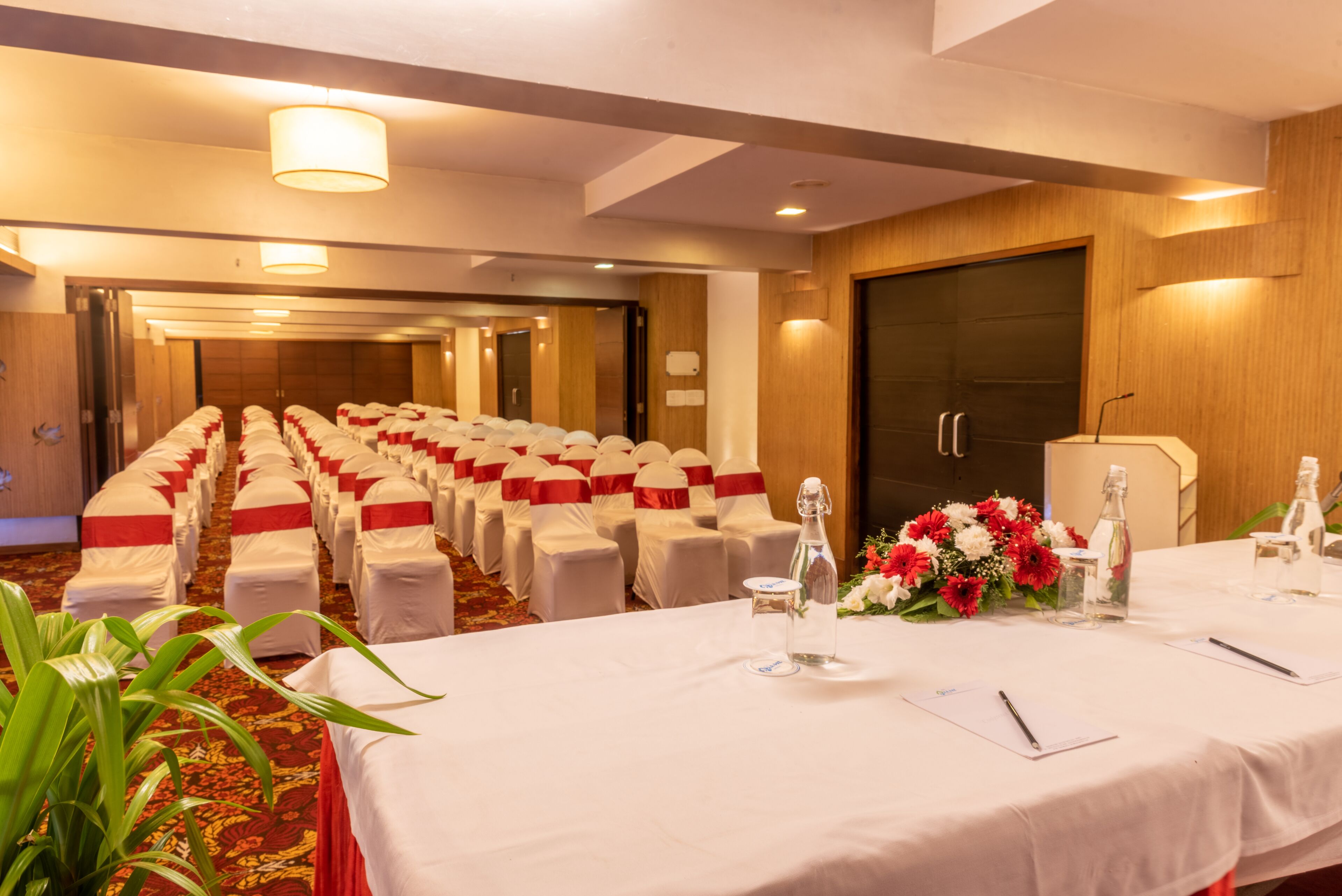 Un salón de conferencias con filas de sillas blancas, una mesa larga con botellas de agua y bolígrafos, y una puerta de madera al fondo.