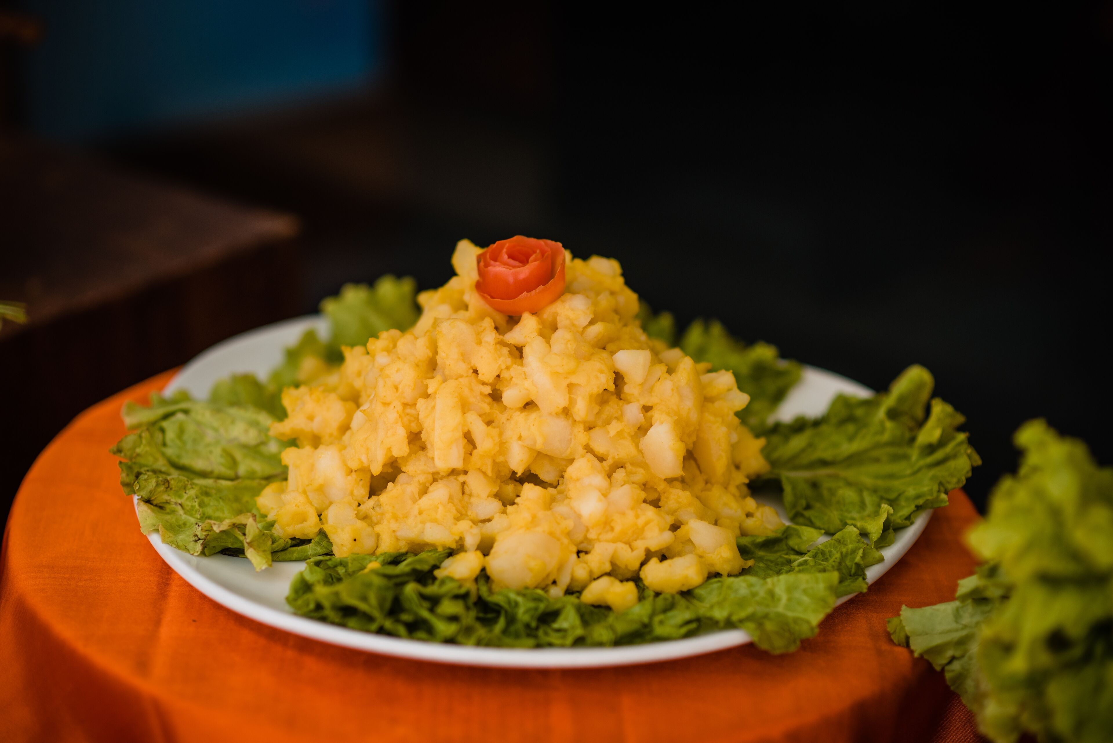 Un plato de comida con un tomate decorativo encima, rodeado de hojas de lechuga, sobre un mantel naranja.