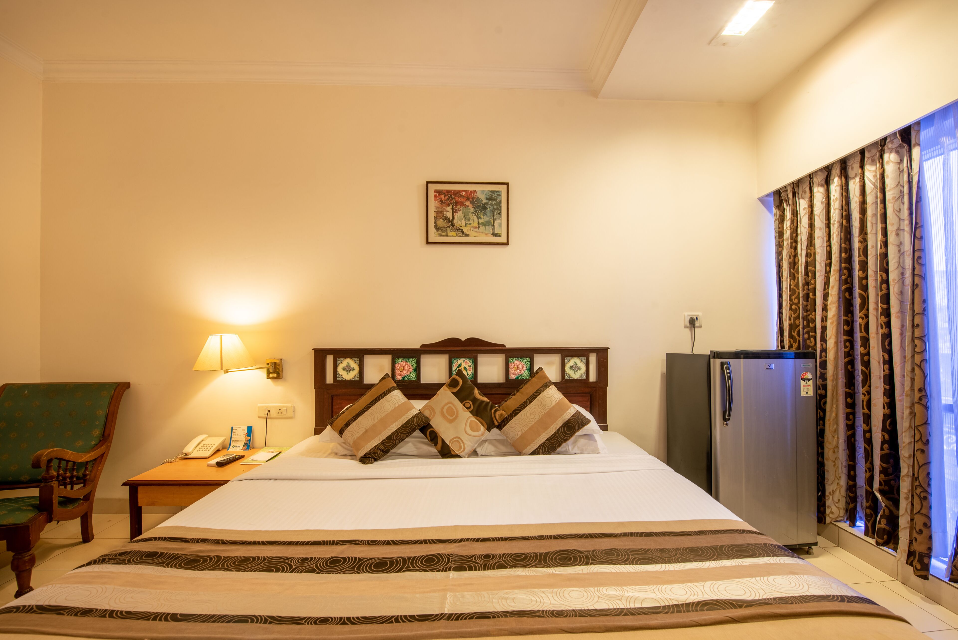 A hotel room with a bed, a nightstand, a lamp, a chair, a refrigerator, and a window with curtains.
