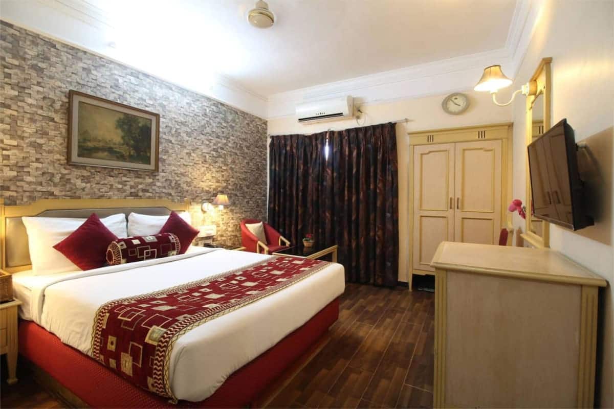 A hotel room with a large bed, a desk, a chair, a TV, a clock, and a painting on the wall.