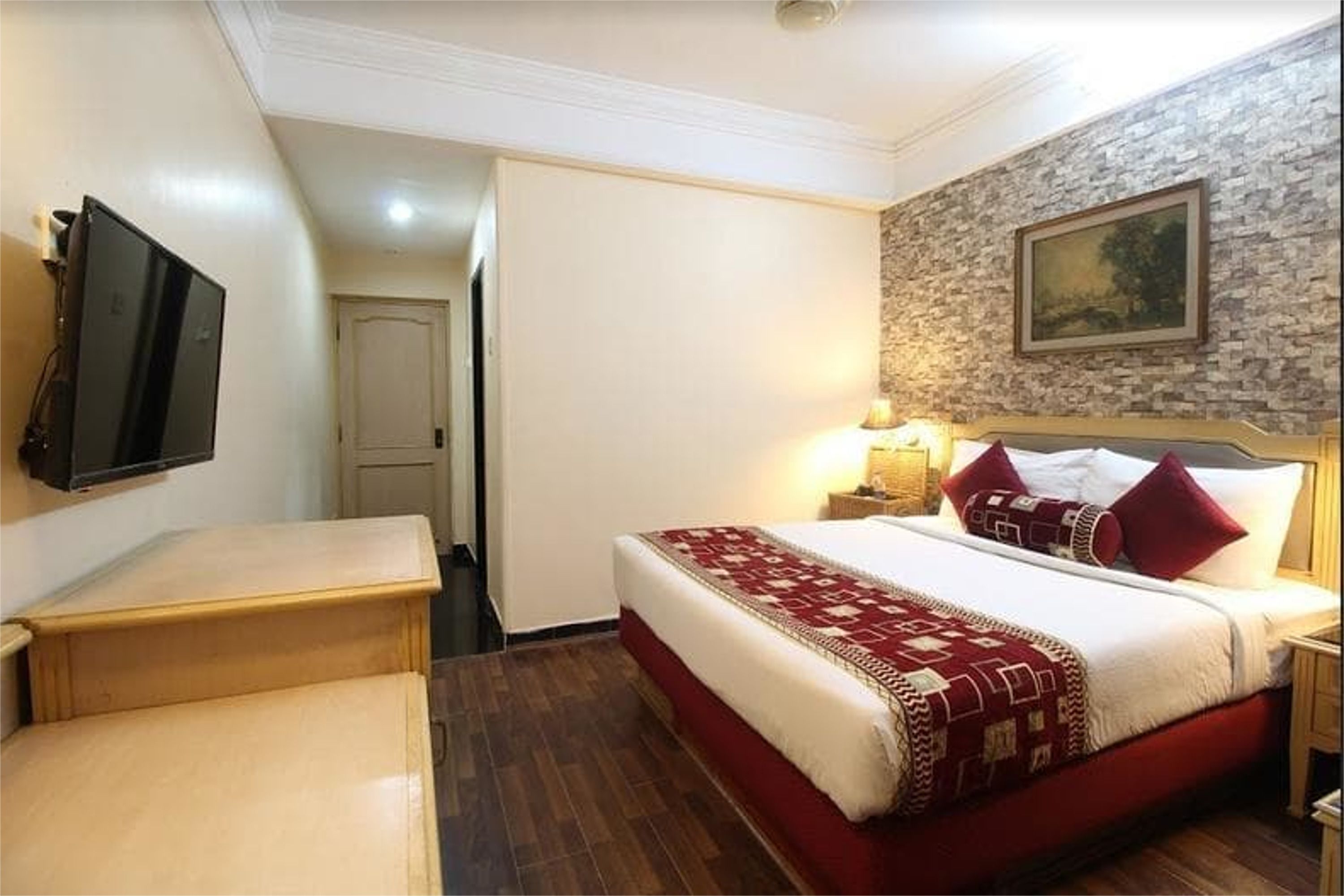 A hotel room with a bed, a TV mounted on the wall, a wooden bench, and a stone wall with a painting.