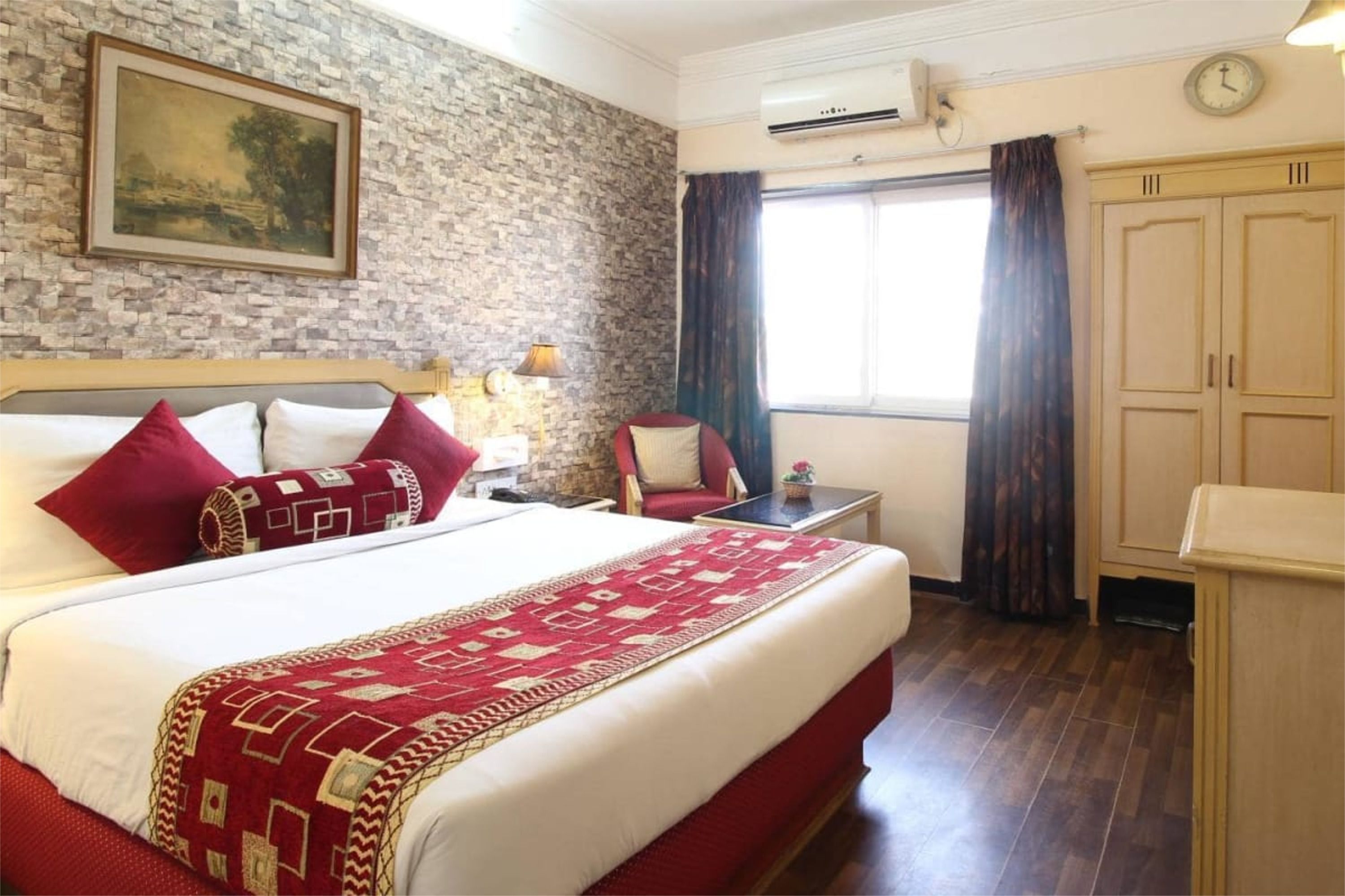 A hotel room with a large bed, a wooden headboard, a stone wall, a painting, a window with curtains, a desk, and a wardrobe.