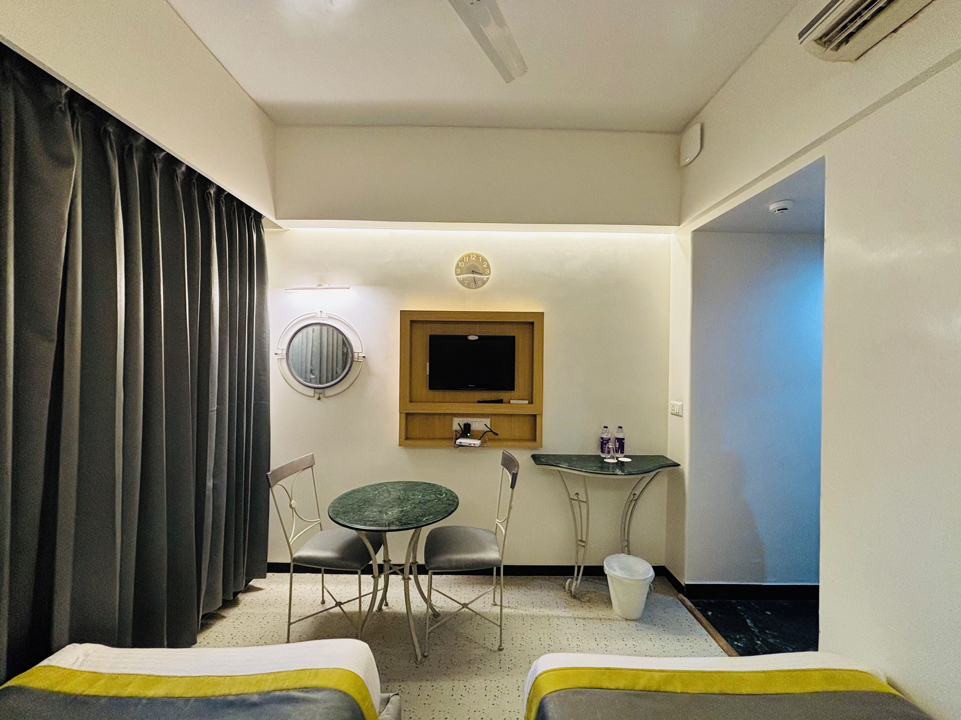 A hotel room with two beds, a round mirror, a television, a small table with chairs, and a window with curtains.