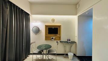 A hotel room with two beds, a round mirror, a television, a small table with chairs, and a window with curtains.