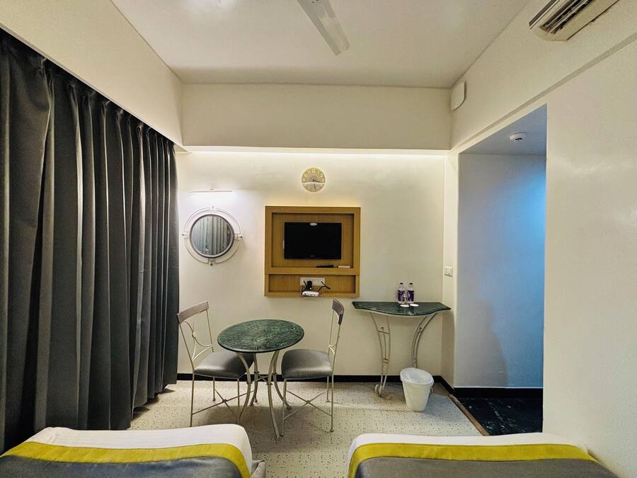 A hotel room with two beds, a round mirror, a television, a small table with chairs, and a window with curtains.