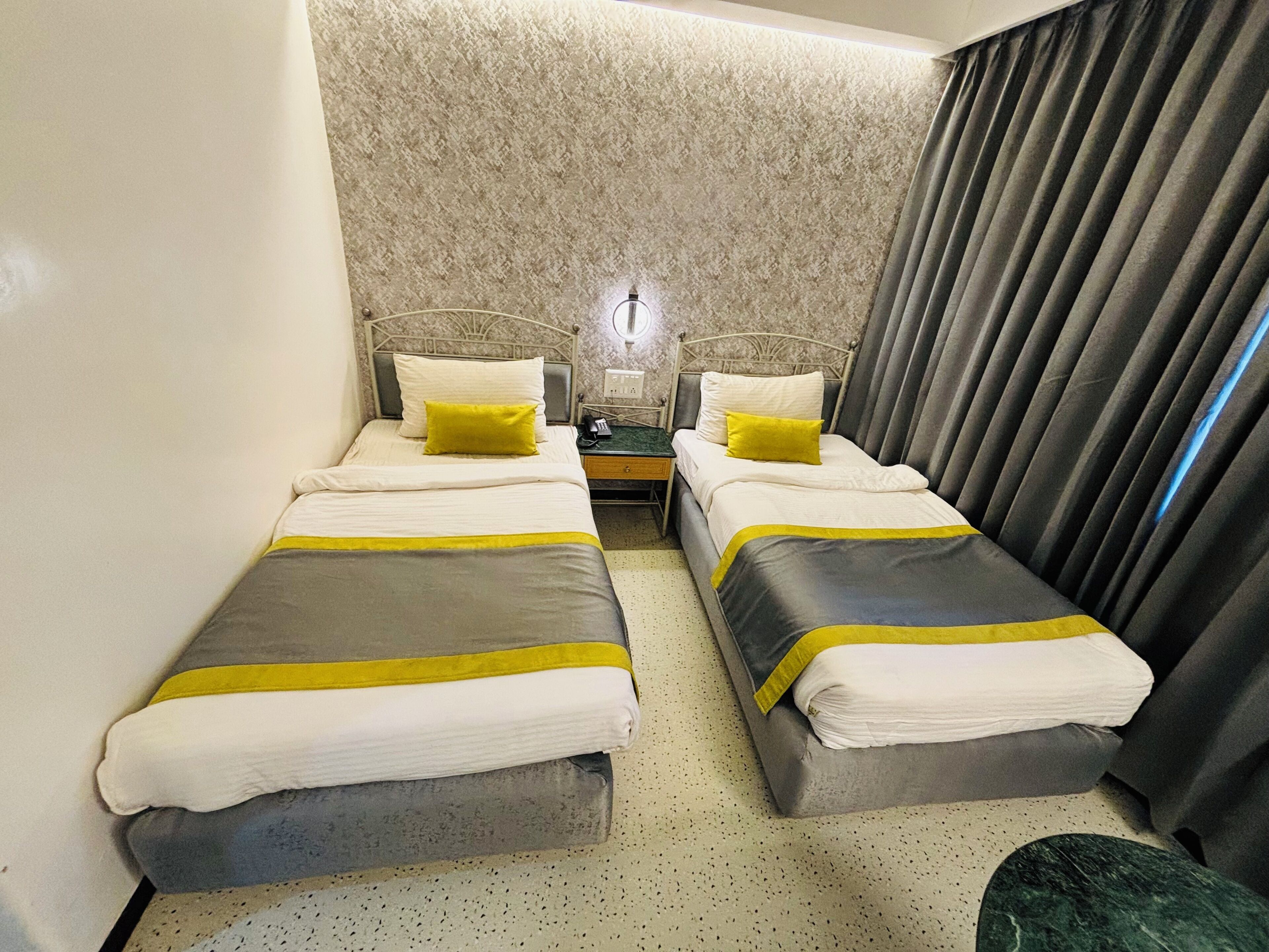 Habitación de hotel con dos camas individuales, cada una con cubrecama gris y blanco, un cojín de color amarillo y una mesita de noche con lámpara.