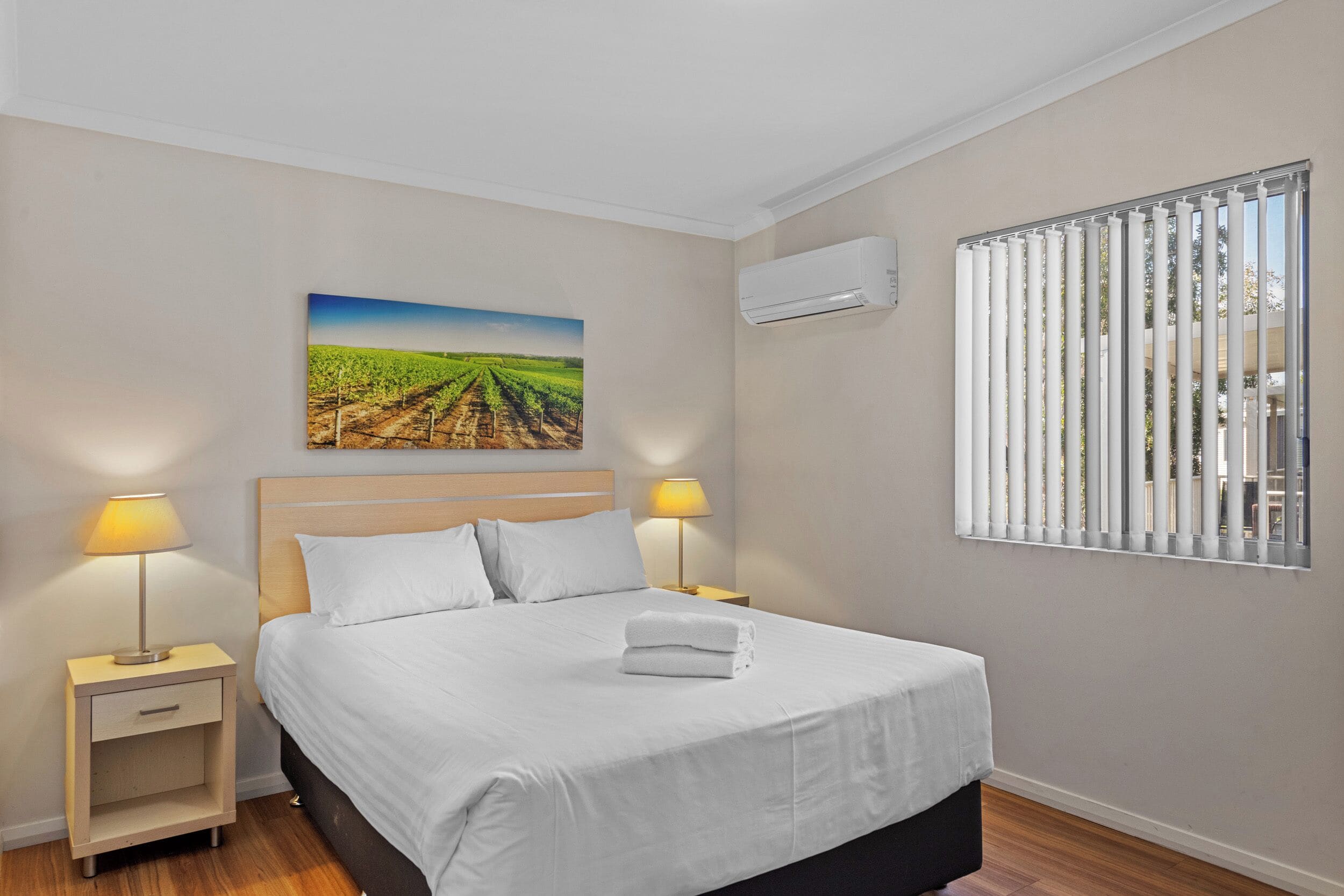 A neatly made bed with white linens, two bedside lamps, a nightstand, a wall-mounted air conditioner, and a window with vertical blinds.