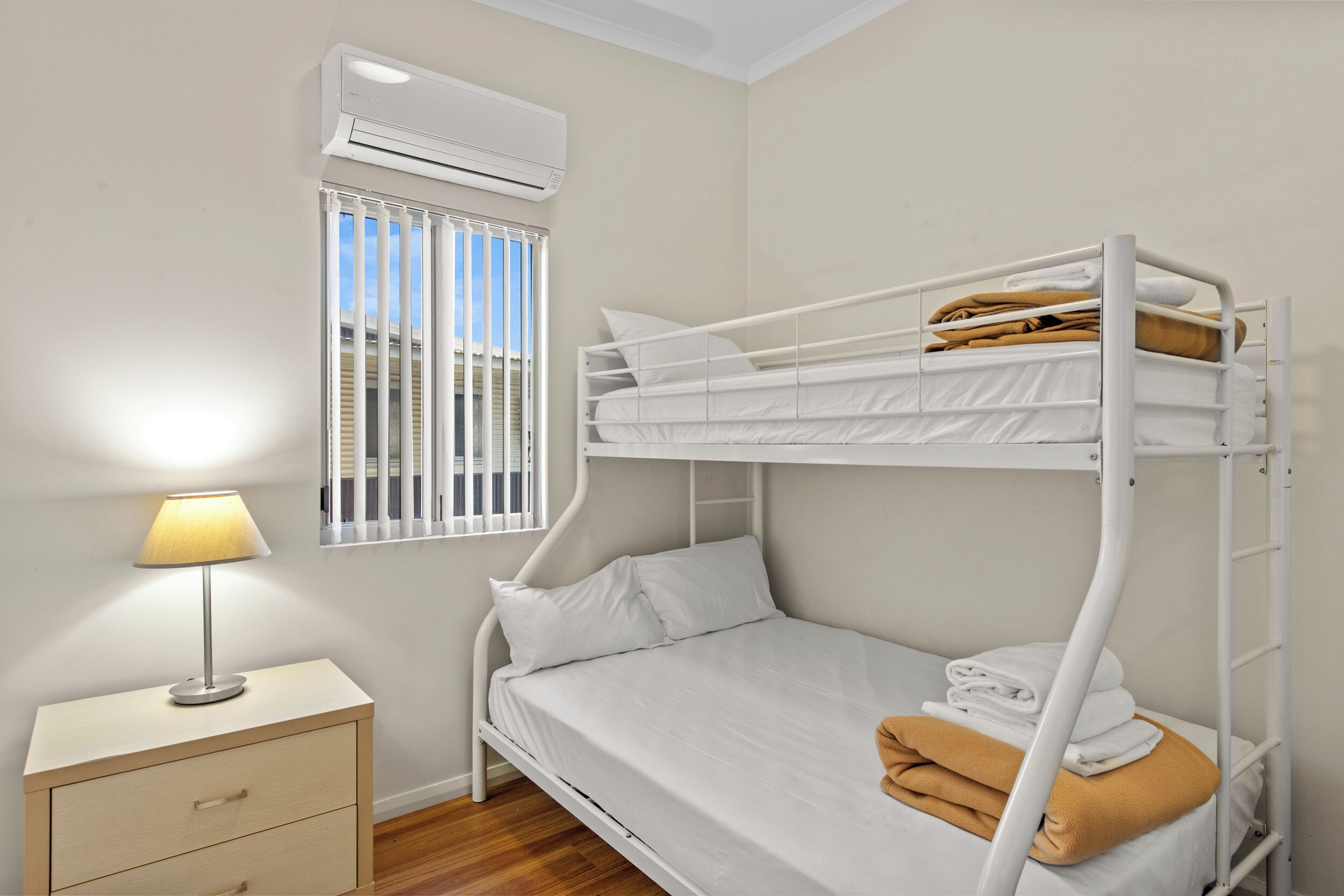 Una cama litera con ropa blanca, una mesita de noche con una lámpara y una ventana con persianas.