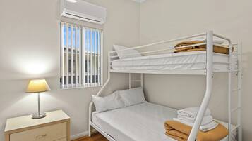 Una cama litera con ropa blanca, una mesita de noche con una lámpara y una ventana con persianas.