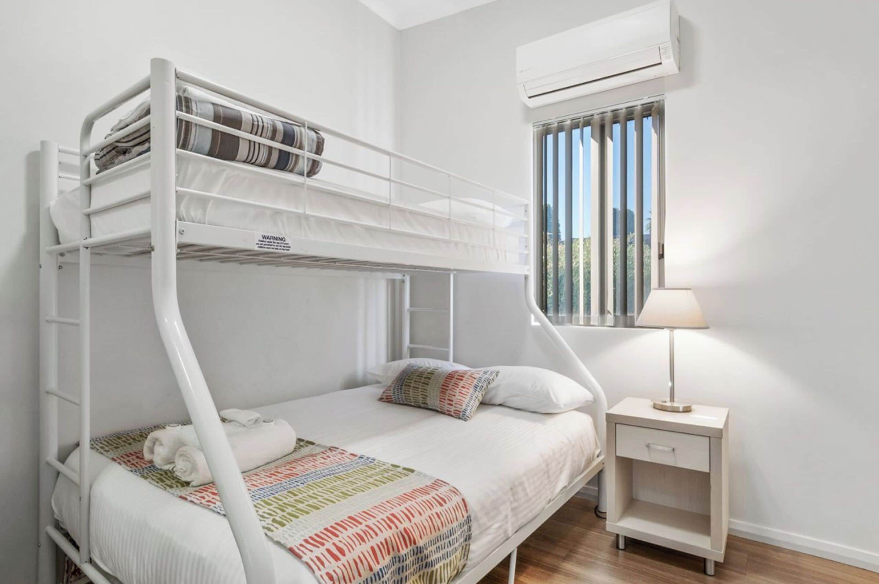Une chambre avec un lit superposé, décorée dans des tons de blanc, comprenant une petite table de chevet avec une lampe et une fenêtre équipée de stores.