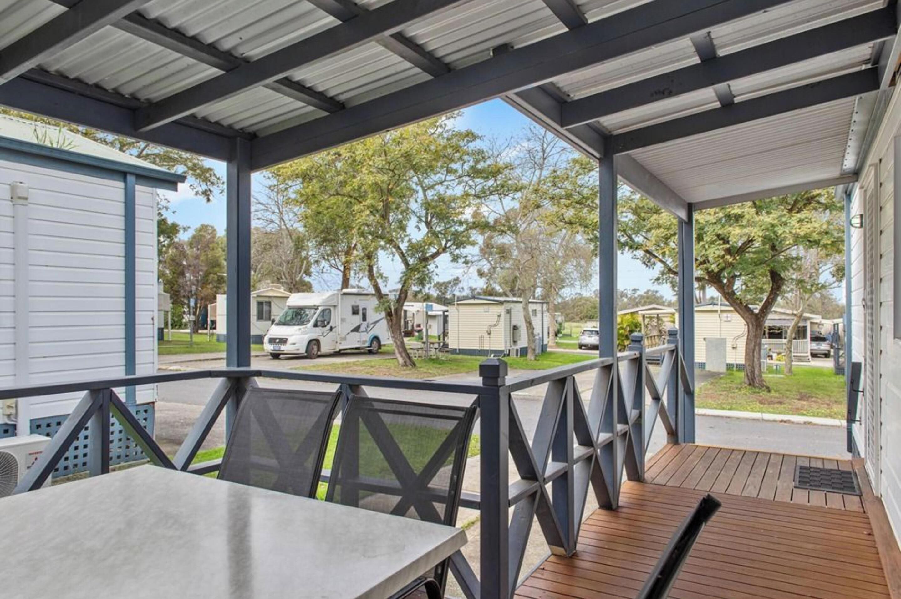 A covered outdoor area with a wooden deck, metal railing, and a view of mobile homes and trees in the background.