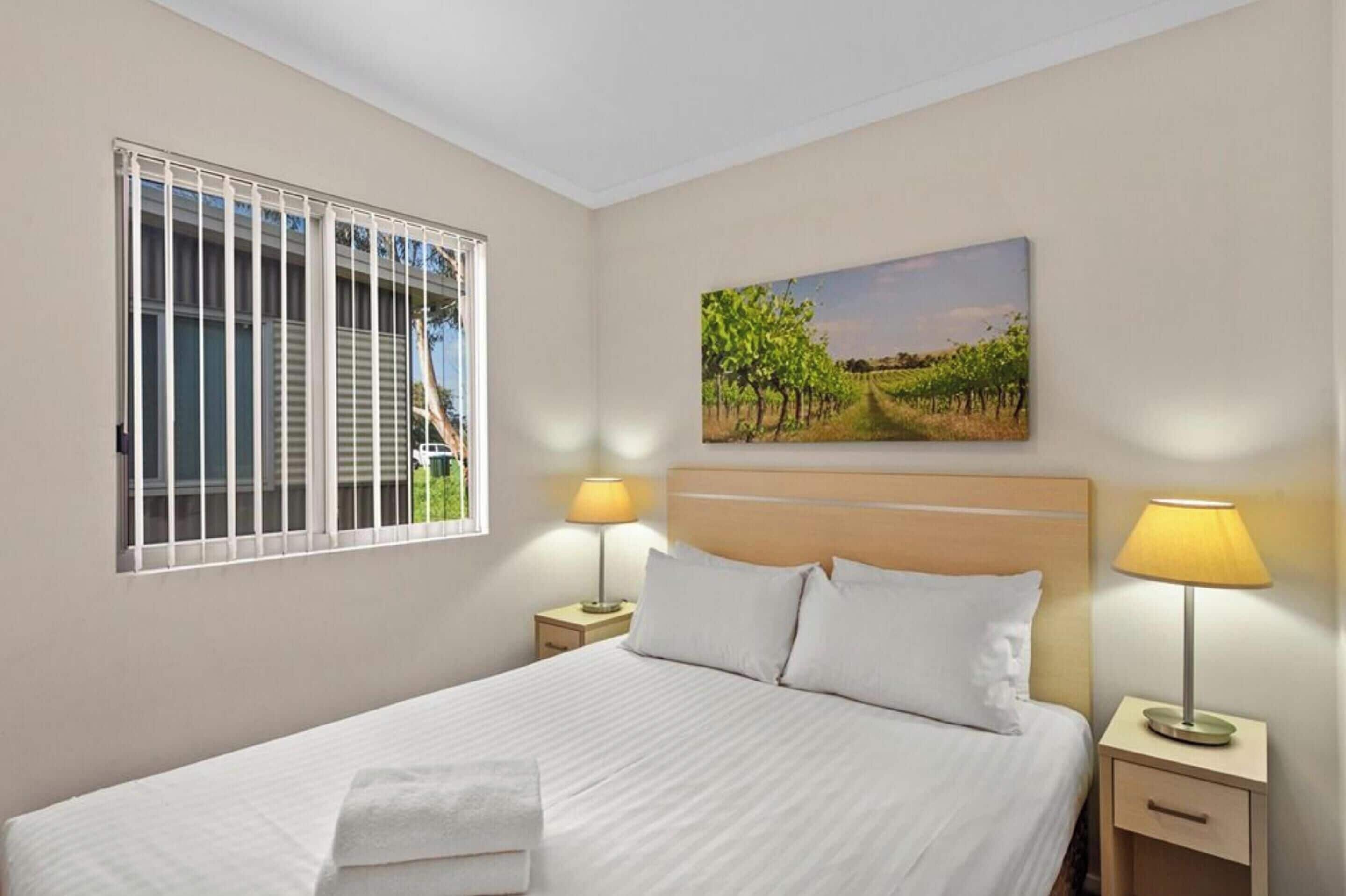 A hotel room with a bed, two bedside tables with lamps, a window with blinds, and a painting of a vineyard on the wall.