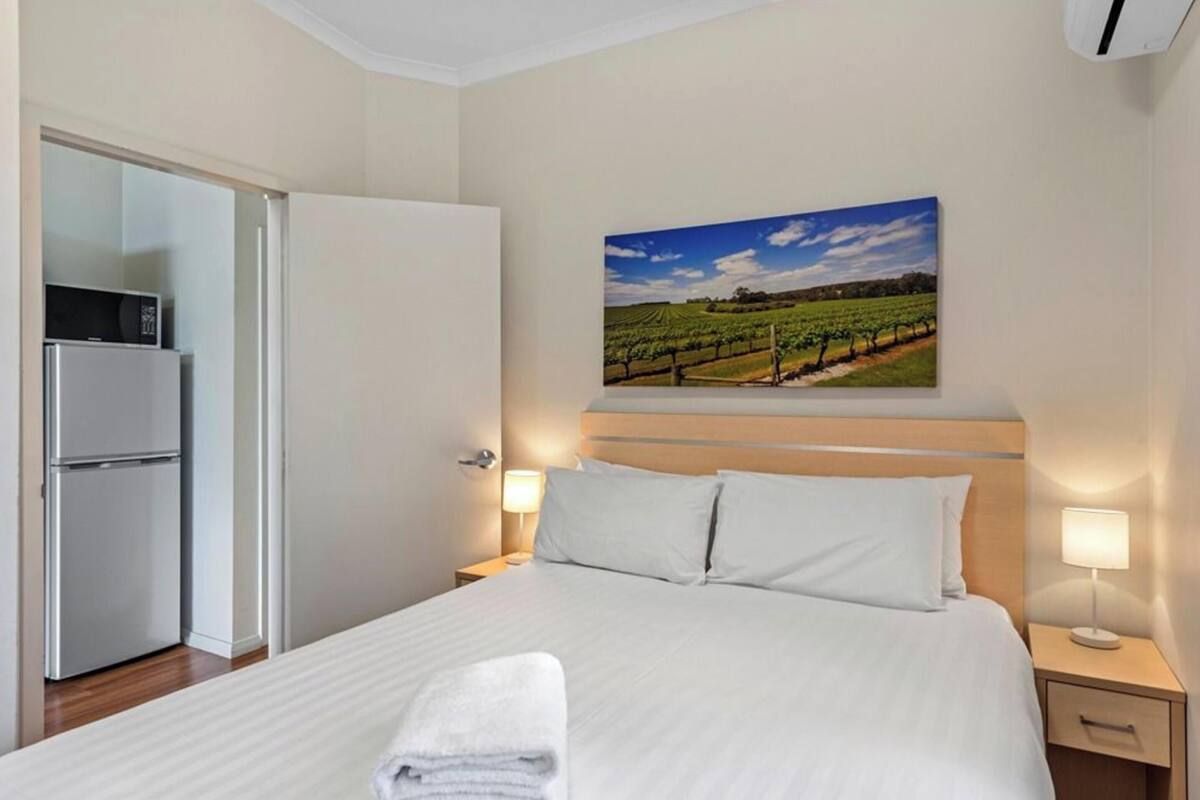 Habitación de hotel con cama, mesita de noche con lámpara, cuadro colgado en la pared de un viñedo, y puerta que da a una kitchenette con refrigerador.