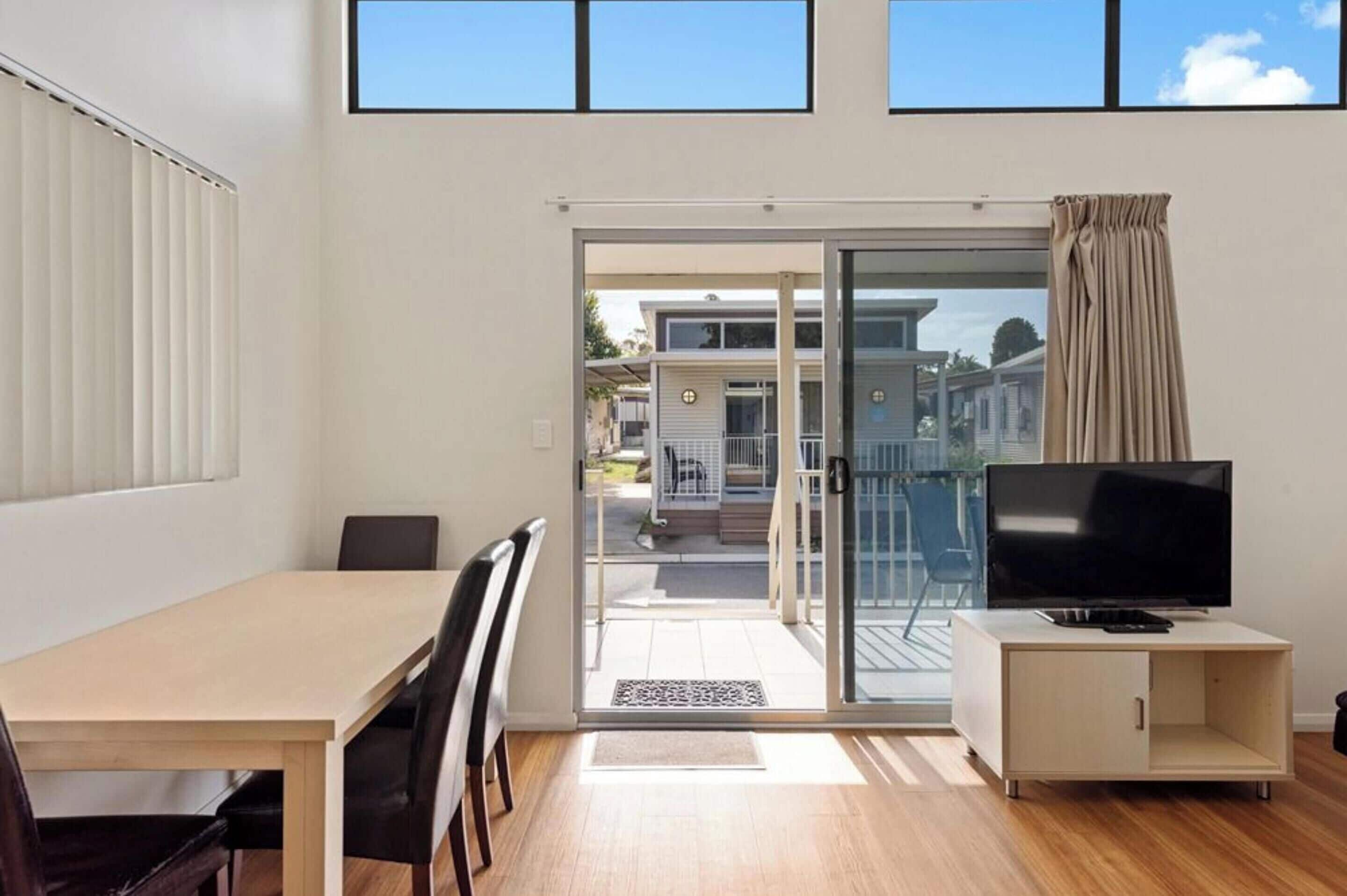 Un salon moderne doté d’une porte-fenêtre coulissante donnant sur un balcon, d’un téléviseur à écran plat posé sur un meuble et d’un coin repas avec une table et des chaises.