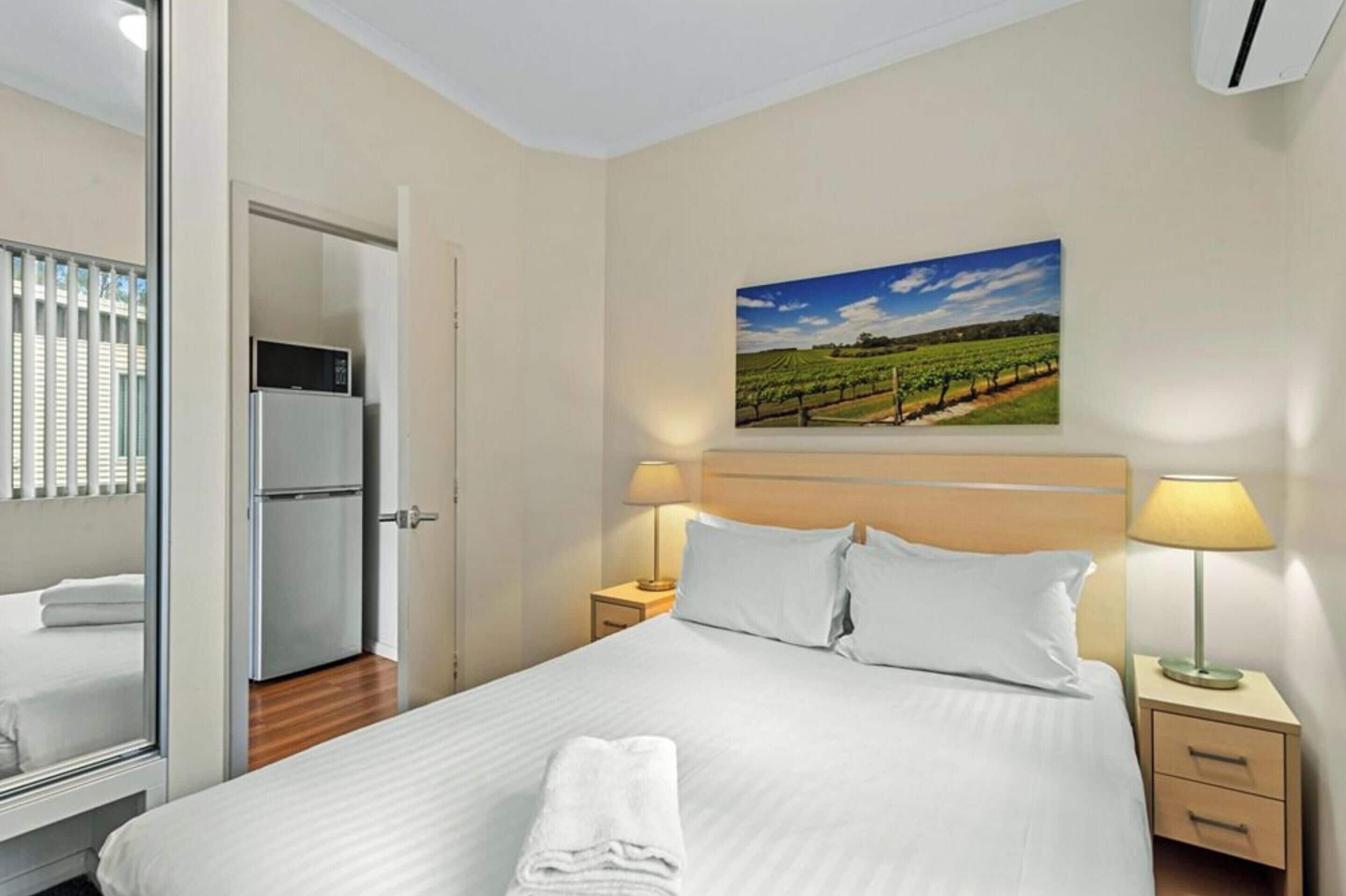 A hotel room with a bed, a refrigerator, a bedside table with a lamp, and a painting of a vineyard on the wall.