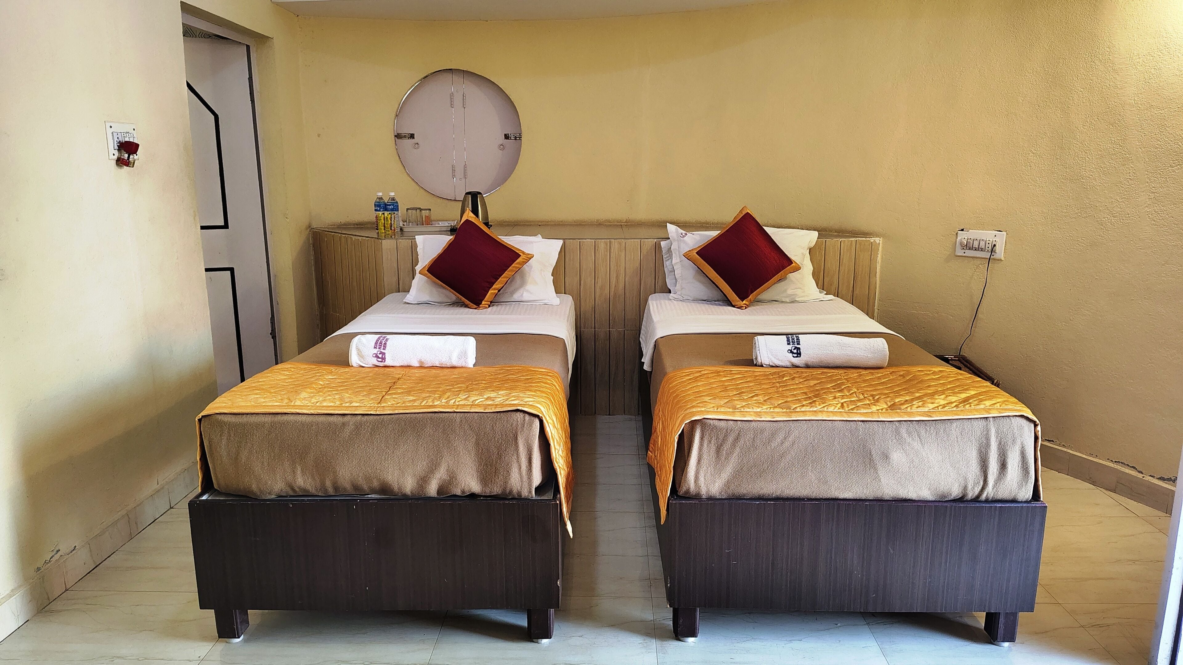 A hotel room with two single beds, a round mirror on the wall, a small table with toiletries, and a door with a light switch.