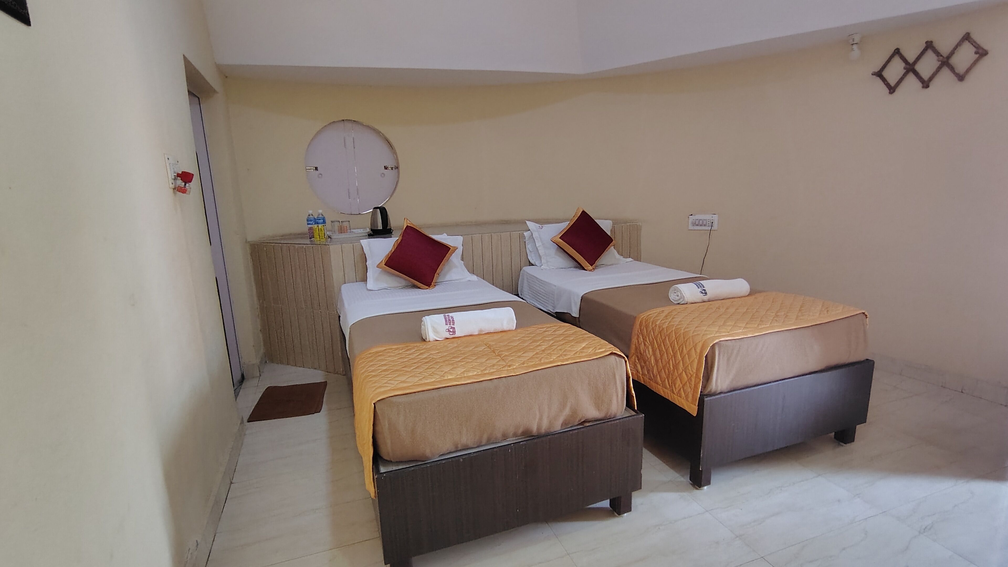 A hotel room with two beds, each with a brown bedspread and a red pillow. There is a small table with a water bottle and a cup, and a wall-mounted light fixture.