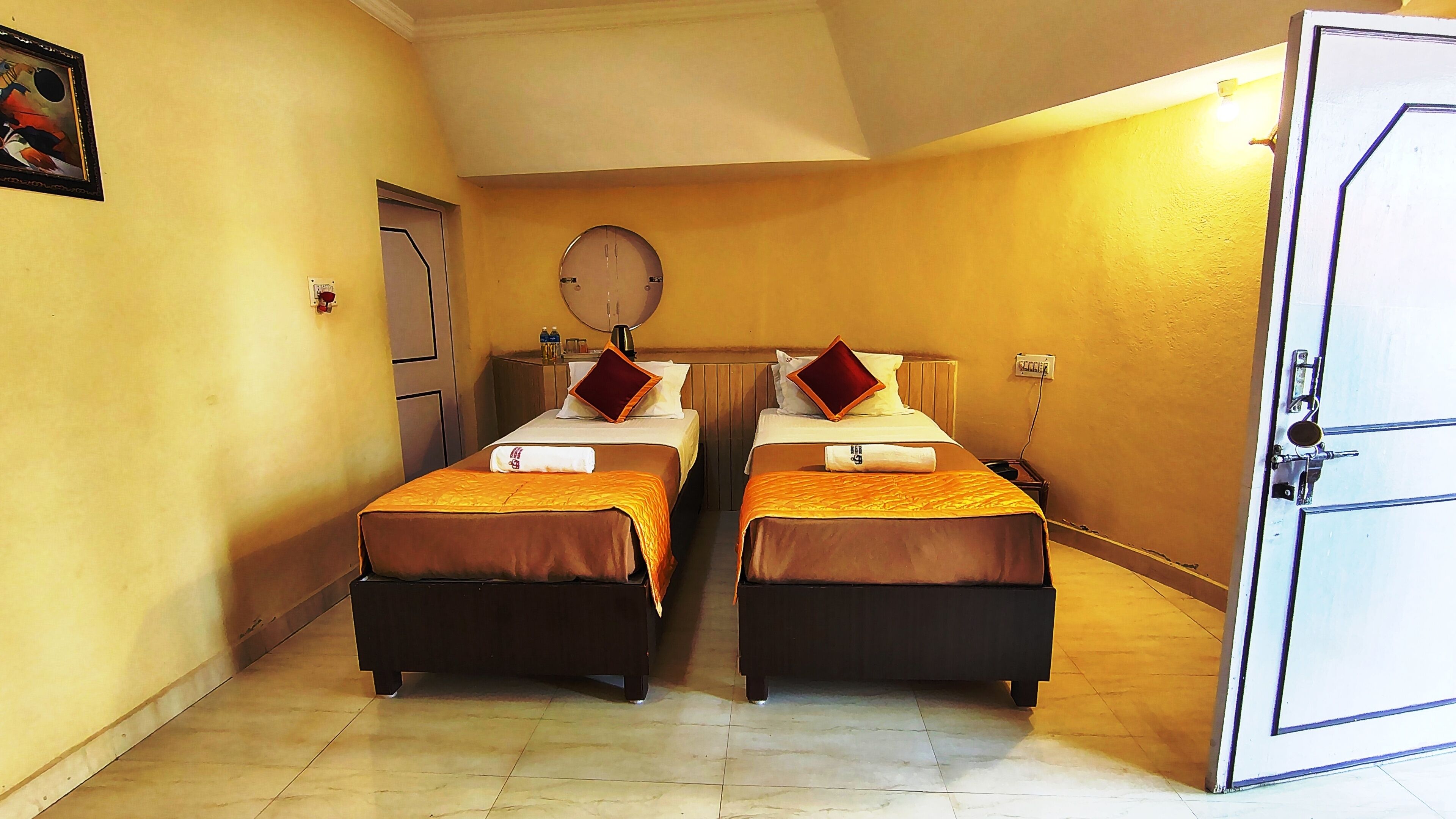 A room with two beds, a door, a wall clock, and a small table with a lamp.