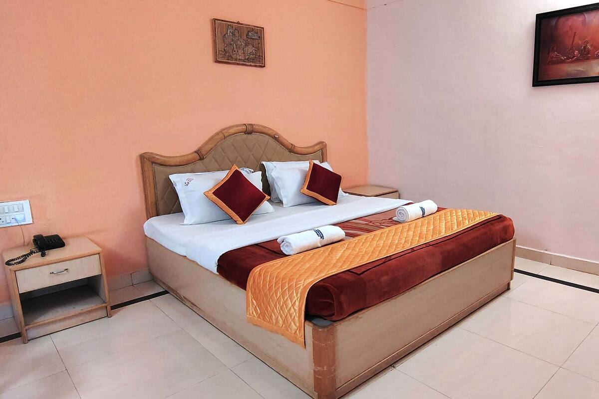 A neatly made bed with a wooden headboard, white linens, and a red throw blanket. There is a small bedside table with a phone and a power outlet.
