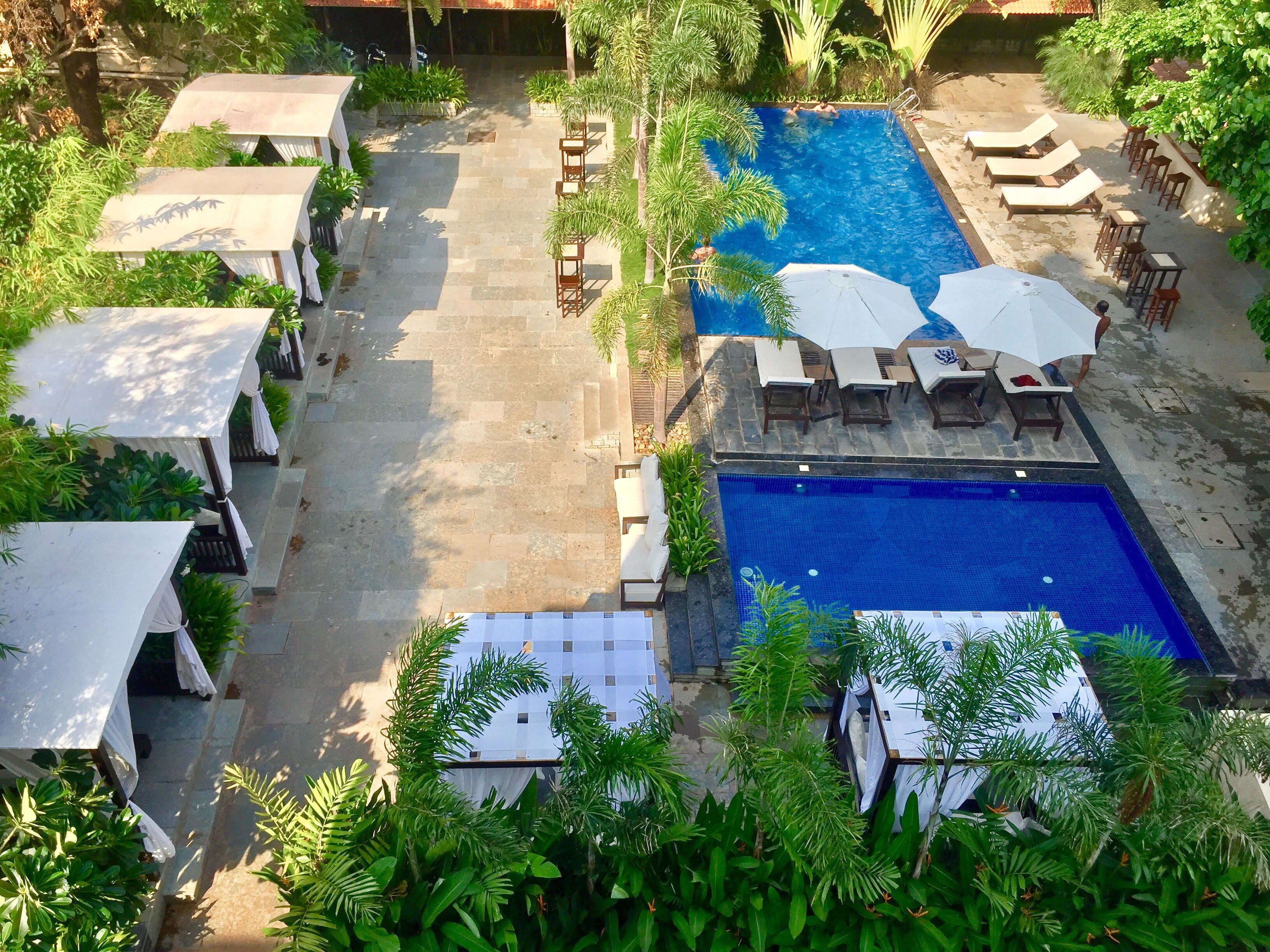 A rooftop pool area with lounge chairs and umbrellas, surrounded by greenery and covered structures.